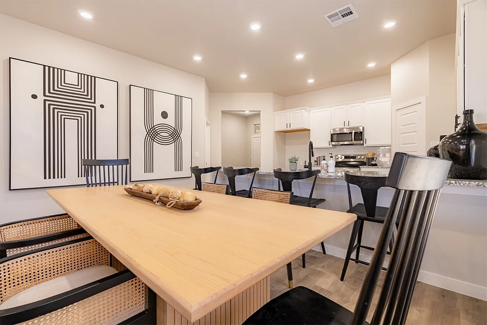 Interior of a modern kitchen and dining area with a wooden table, black and beige chairs, white cabinets, stainless steel appliances, and black and white abstract artwork on the wall.