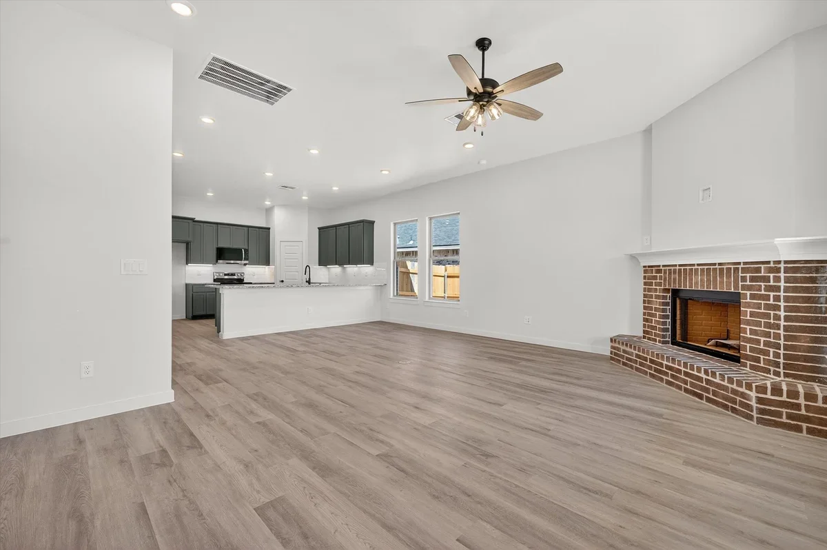 Empty living room with hardwood floors, white walls, a brick fireplace, a ceiling fan, and an open kitchen area with gray cabinets and windows.