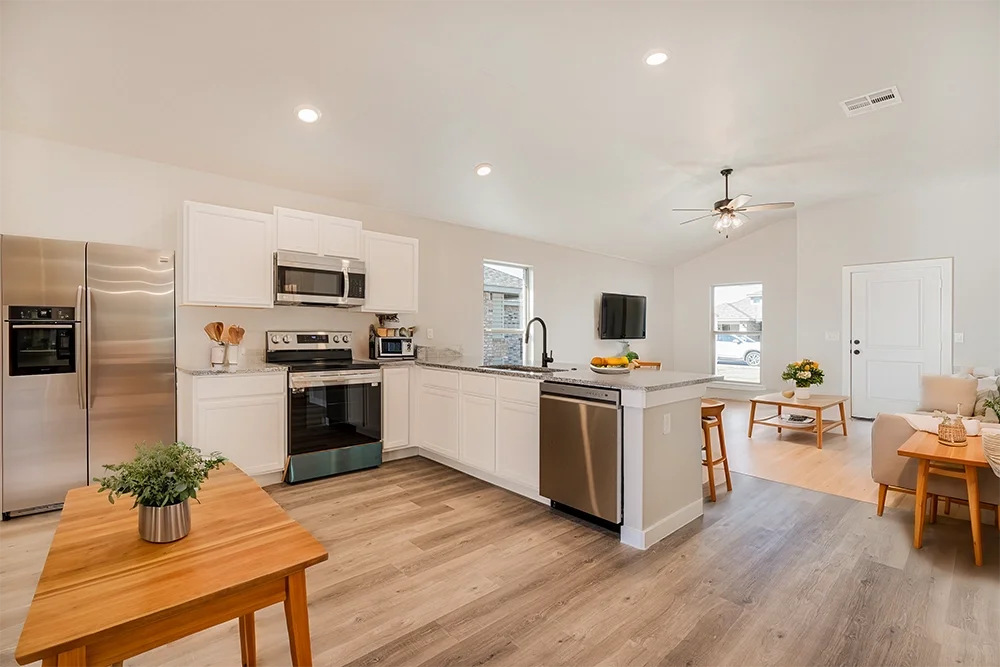 Open-concept kitchen and living room with white cabinets, stainless steel appliances, a granite island, and wooden furniture in a bright space.