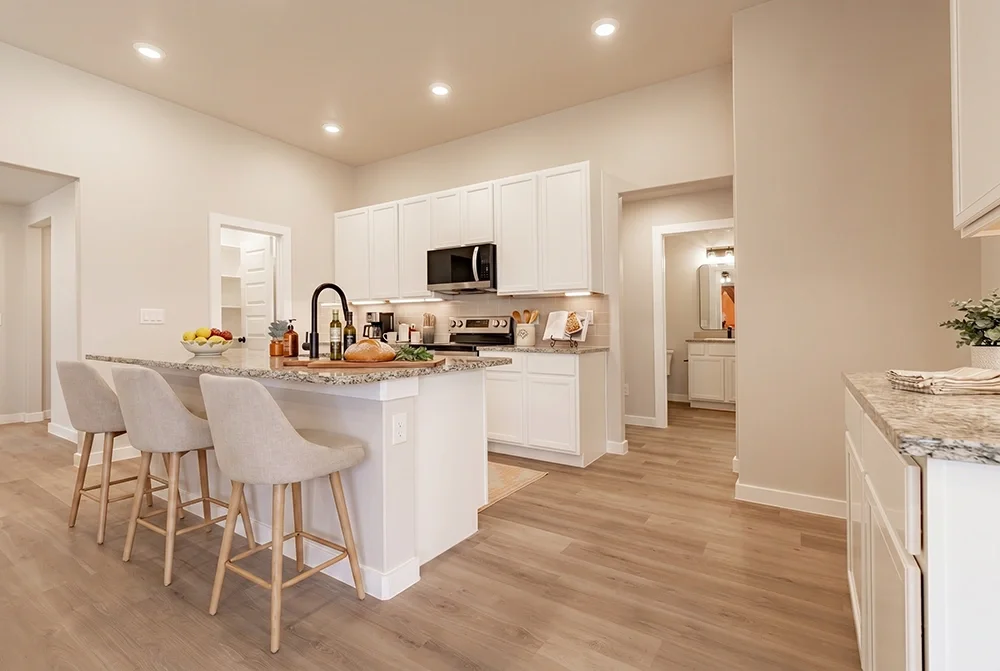 Modern kitchen with white cabinets, granite countertops, island with three upholstered barstools, wood flooring, recessed ceiling lights, and appliances including a microwave and stove.