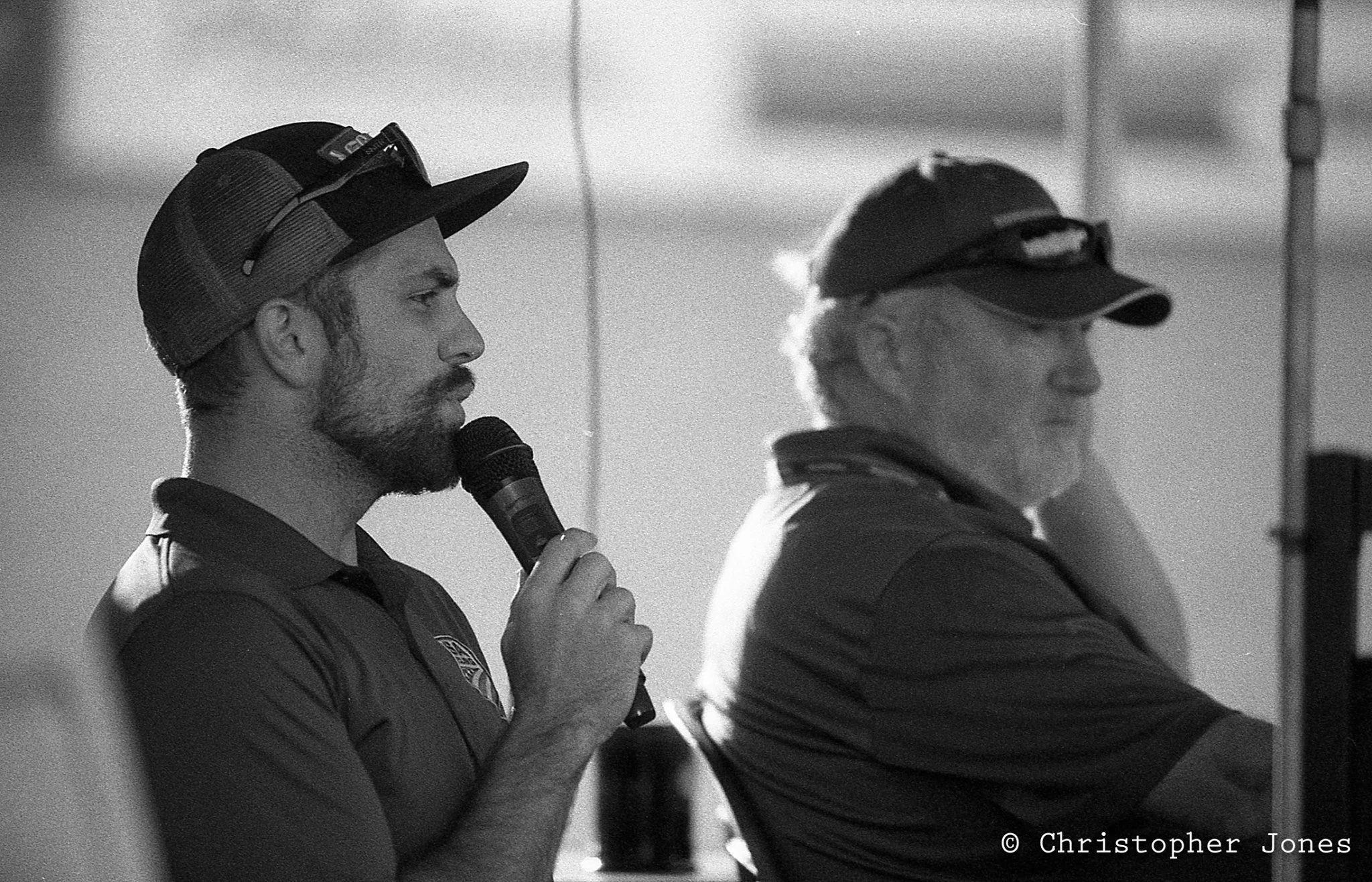Two men in athletic uniforms and caps standing side by side, with one of them holding a microphone. The man with the microphone appears to be speaking or preparing to speak, while the other man looks on. The setting seems to be indoors or in a venue, possibly a sports event or presentation.