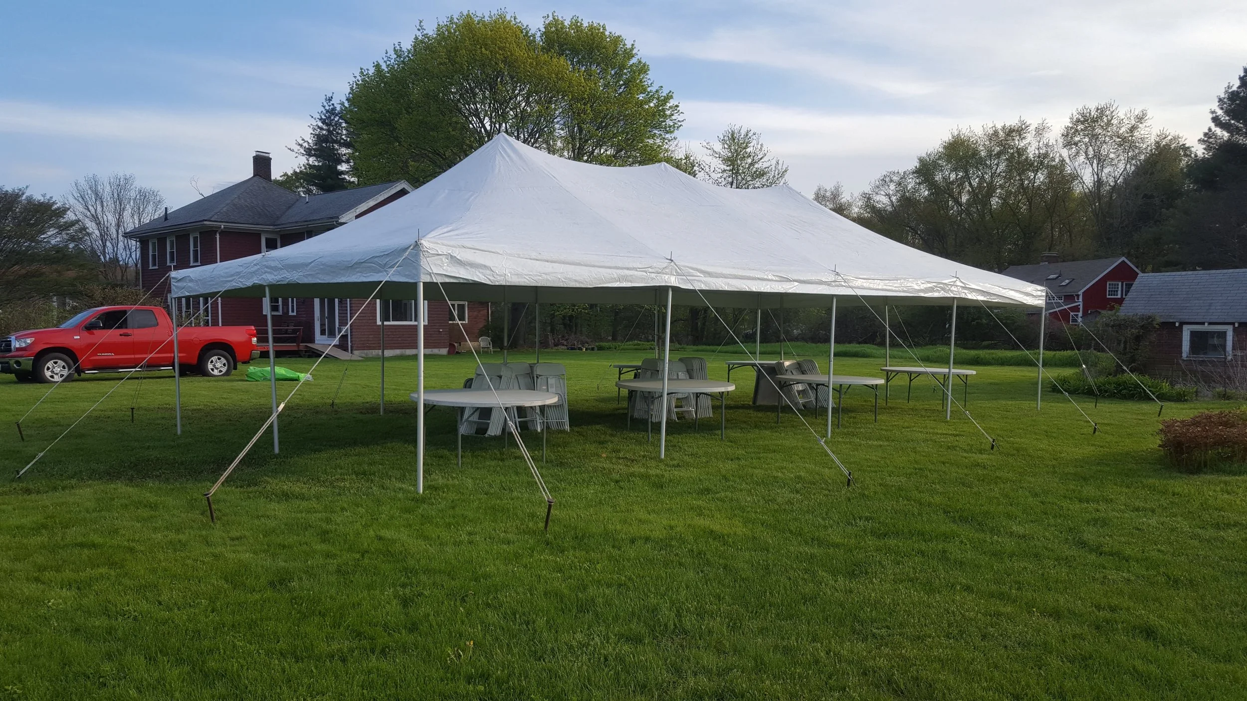 A large white party tent set up on a well-maintained grassy yard, with several plastic tables and chairs underneath, next to a red house with a porch and a red pickup truck parked nearby. In the background, there are trees and additional houses.