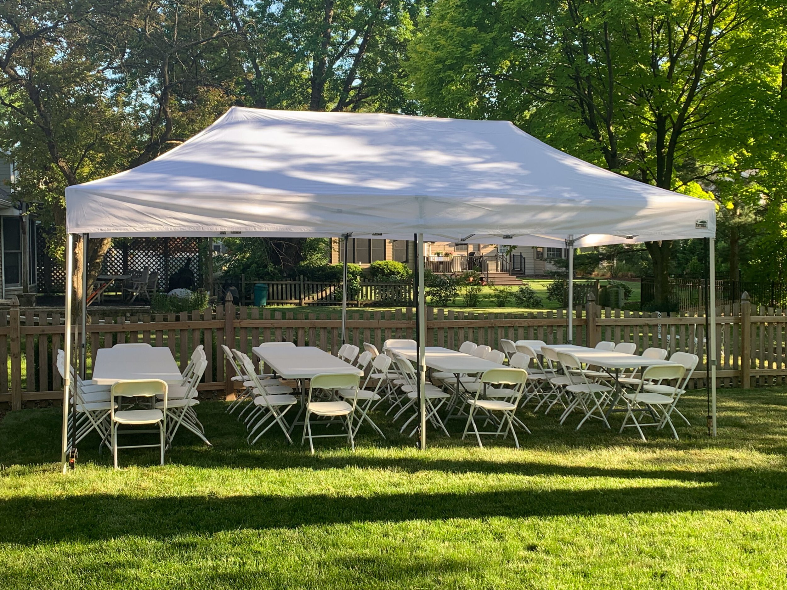 Outdoor setup with white folding chairs and tables arranged under a large white canopy in a backyard, surrounded by trees and a wooden fence.