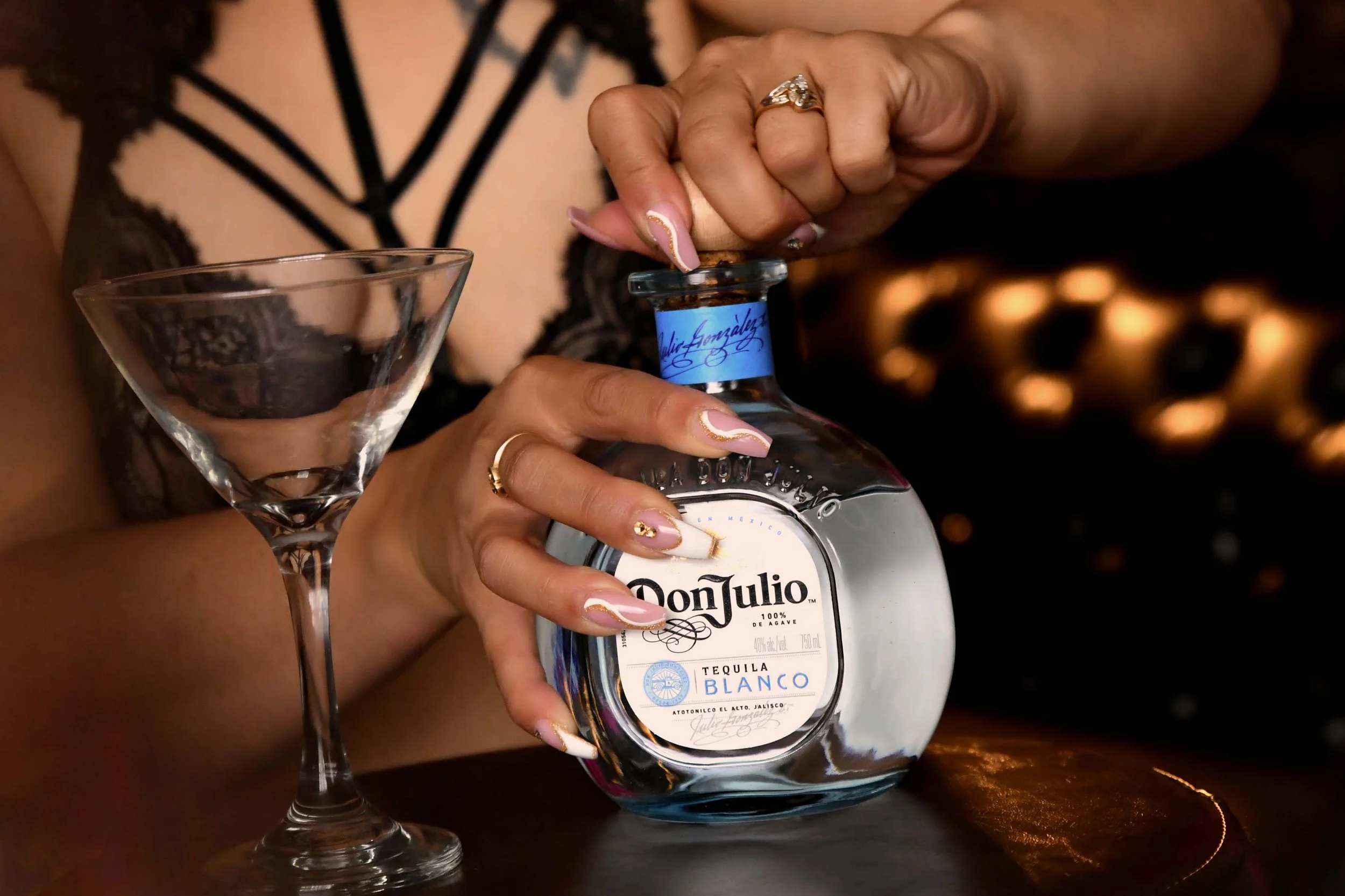  A boudoir detail image of a woman with beautifully manicured nails opening a bottle of tequila with an empty martini glass next to her in Springfield, MO 