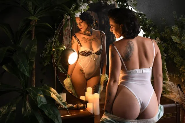 A woman with dark hair wearing white and leopard print lingerie is standing in front of an antique vanity where you can see her reflection as well as her backside