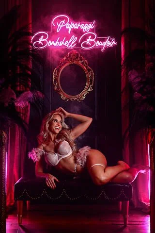 A blonde woman is posed laying on her side on a black leather bench with  a black and neon pink background with the Paparazzi Bombshell Boudoir neon lit up behind her. She is smiling and adjusting her hair for her boudoir shoot near Rogersville, MO