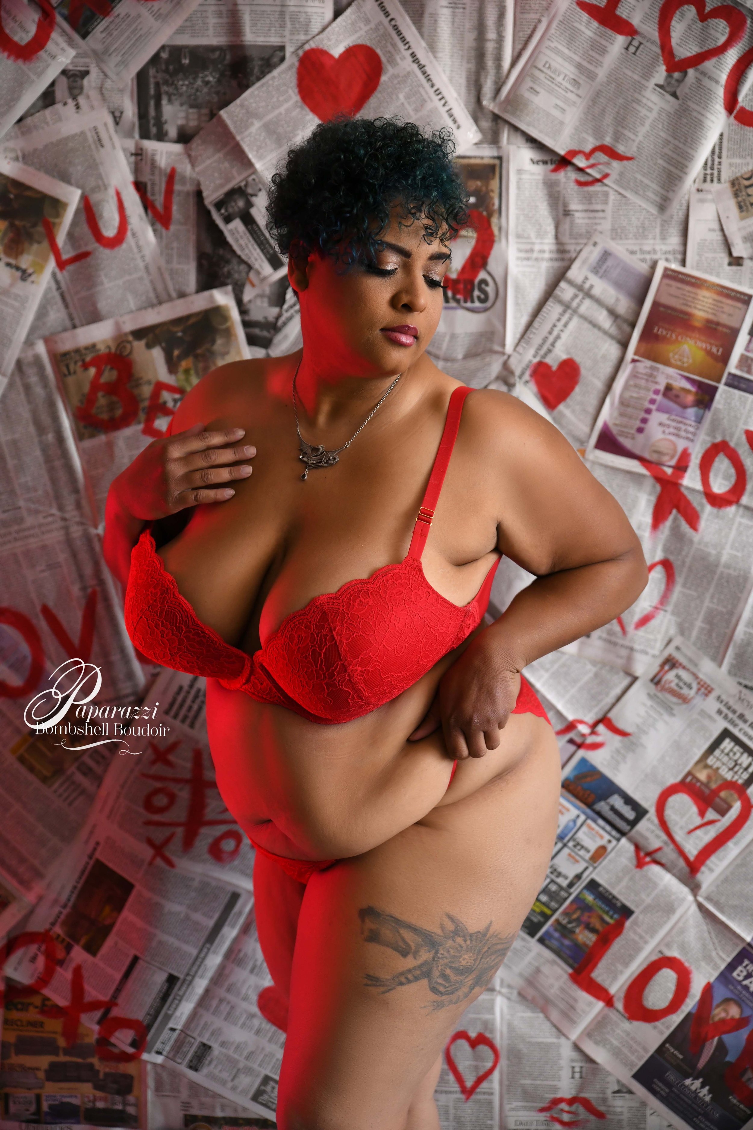 A woman in red lingerie with curly hair, closed eyes, and a tattoo on her thigh, standing amidst newspaper pages decorated with red hearts, lips, and the words 'LOVE' and 'BEAUTY' in a boudoir studio in southwest Missouri