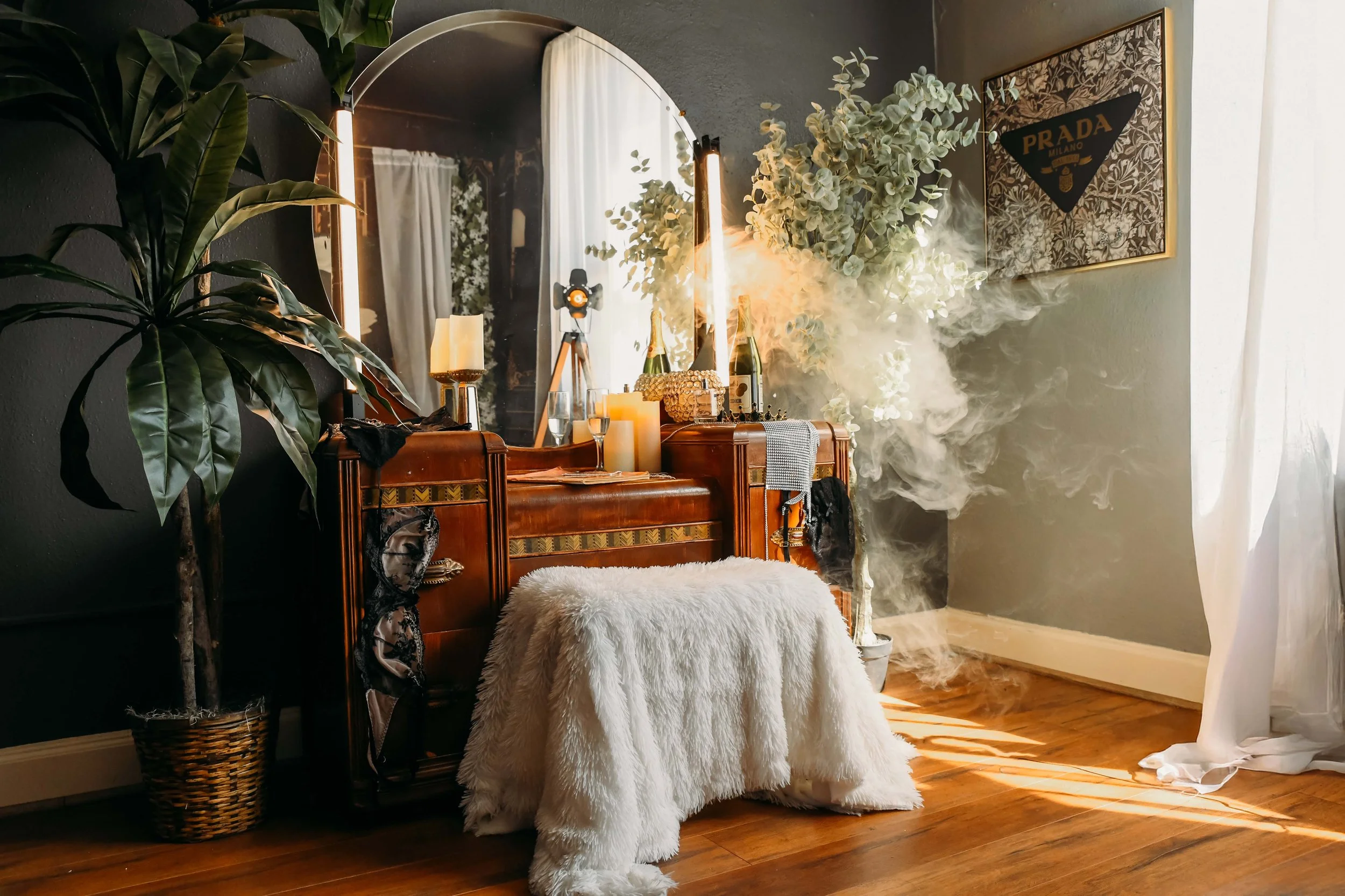 A boudoir set of a vintage wooden vanity with a large mirror and candles, surrounded by green plants and decor. A framed Prada Milano poster hangs on the wall. The room has hardwood floors and a sheer curtain on the right side.