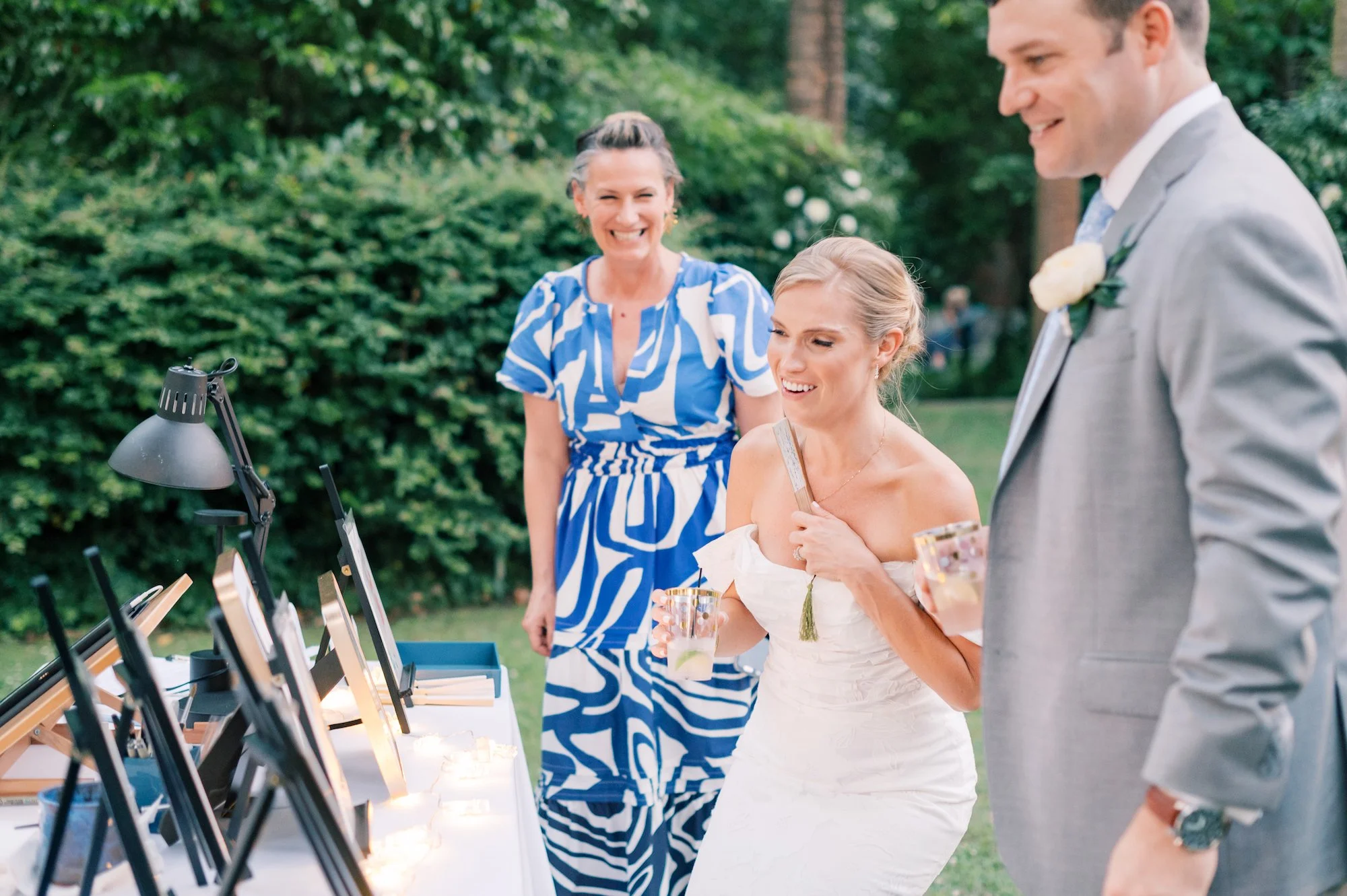 A bride and groom react to the portraits on a table at their wedding with artist, Jamie Peterson smiling on.