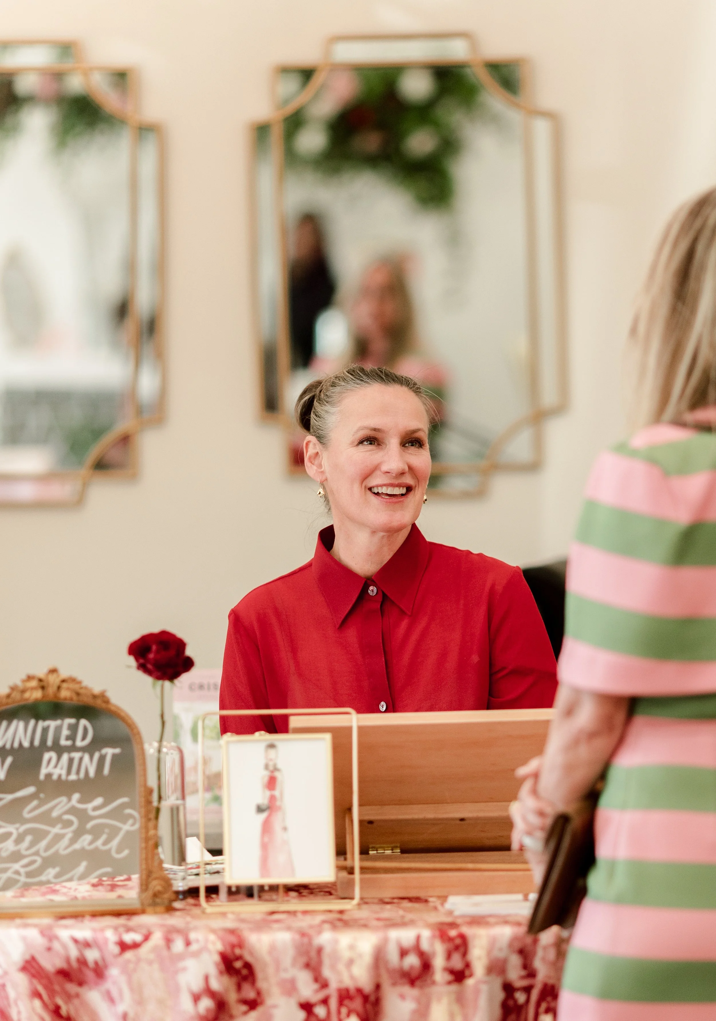 Woman in pink and green stripes approaches a portrait bar table and talks to the artist