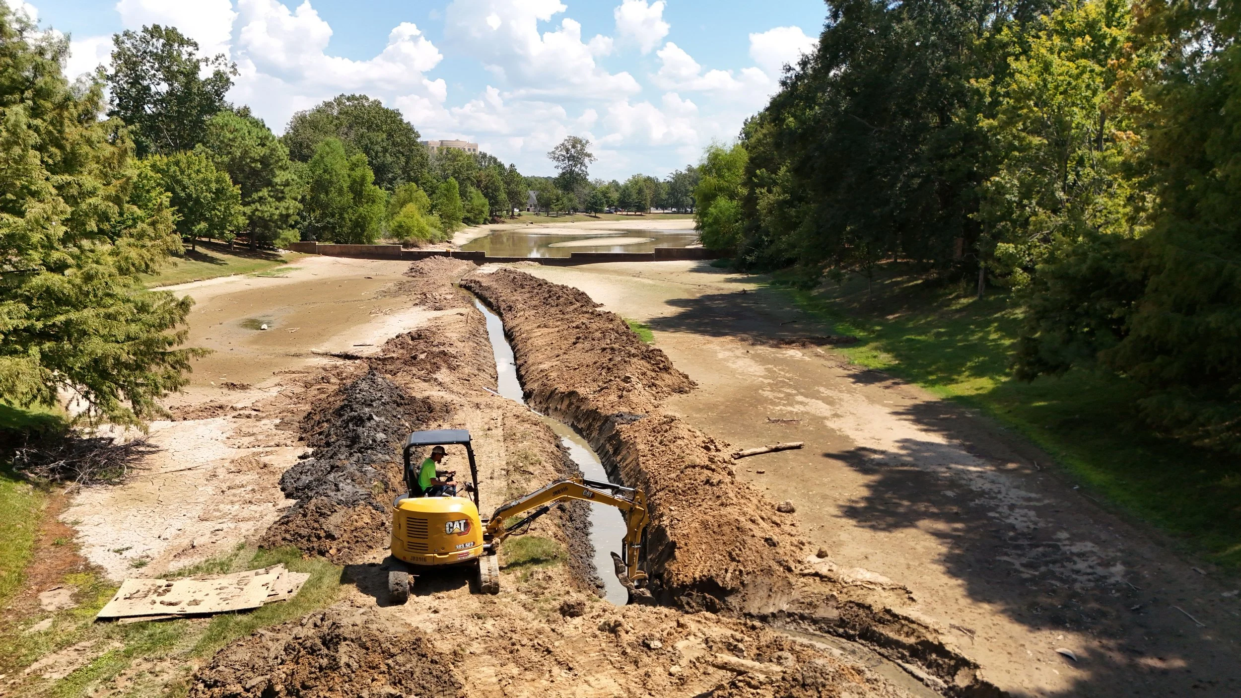 A construction worker operating a small yellow Caterpillar excavator working on a trench near a body of water in a park, with trees and a partly cloudy sky in the background.