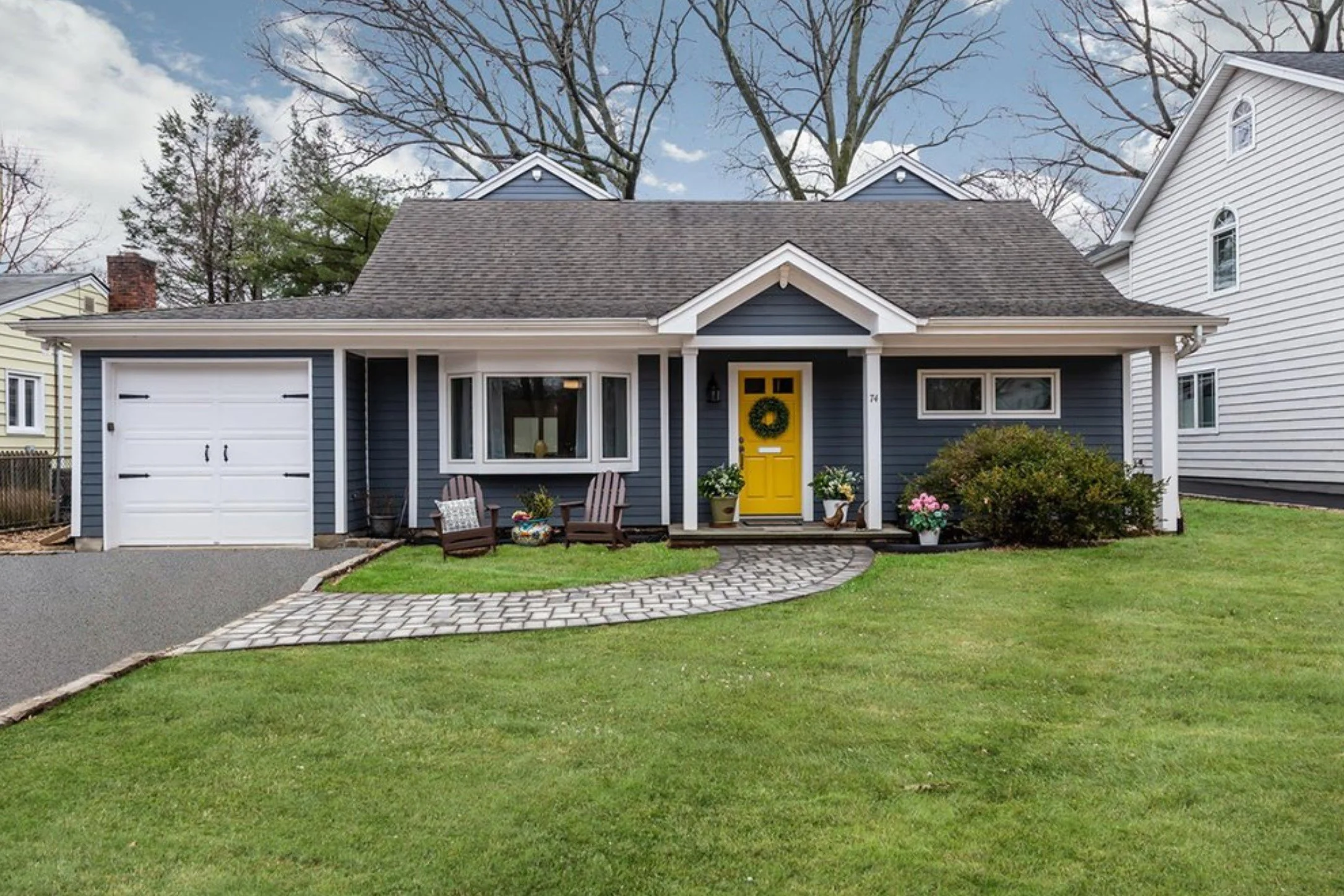 Blue suburban house with a gray roof, yellow front door, white trim, and attached garage. The front yard has a green lawn, potted plants, a stone pathway, and two Adirondack chairs. The sky is partially cloudy.