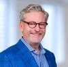 A portrait of a man with glasses, gray hair, and a beard, wearing a blue blazer and checkered shirt, smiling against a neutral background.