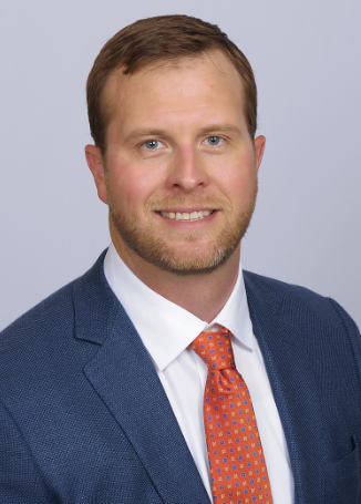 A professional man with short light brown hair, blue eyes, and a beard, wearing a navy blue suit, white shirt, and an orange patterned tie, posed against a plain light gray background.