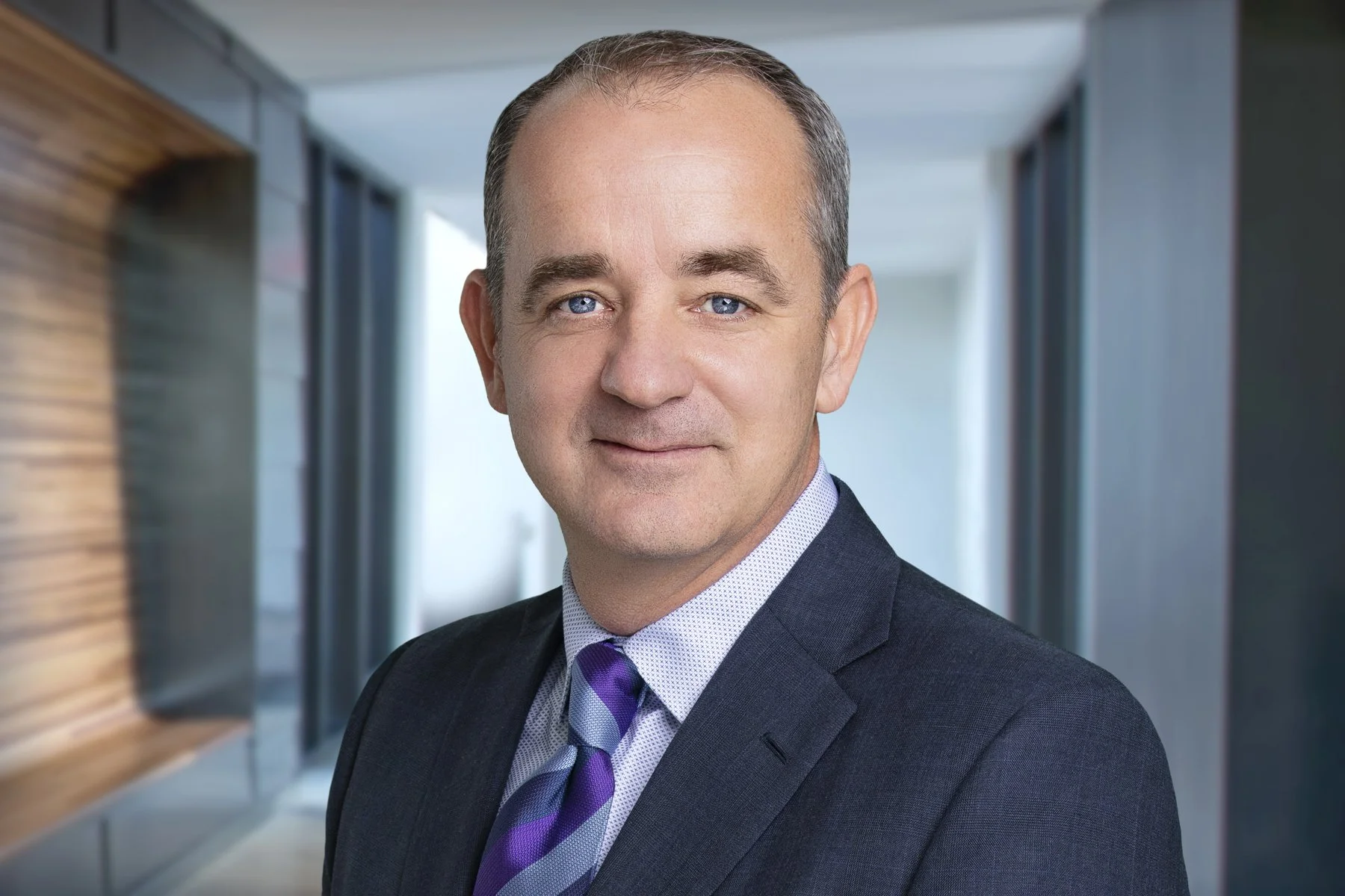 Headshot of a middle-aged man with short gray hair, blue eyes, wearing a dark suit, light shirt, and purple striped tie, standing in a modern office hallway.