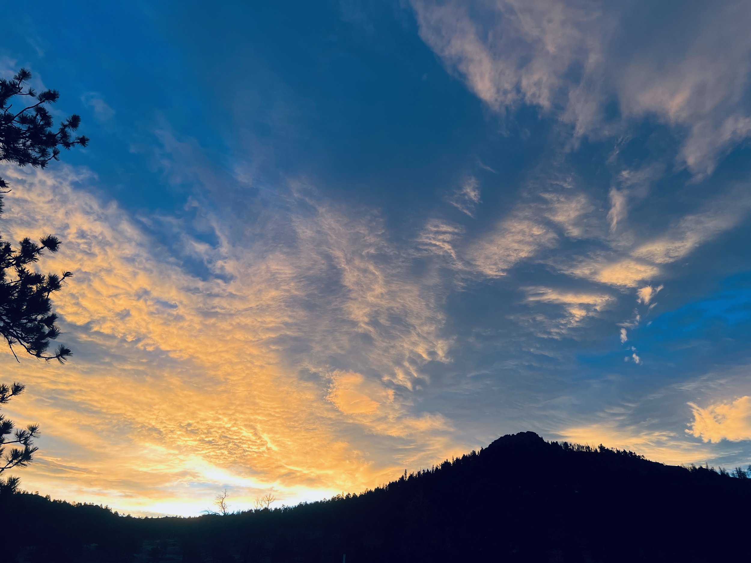 A sunset sky with clouds, silhouette of a mountain, and trees on the left side.