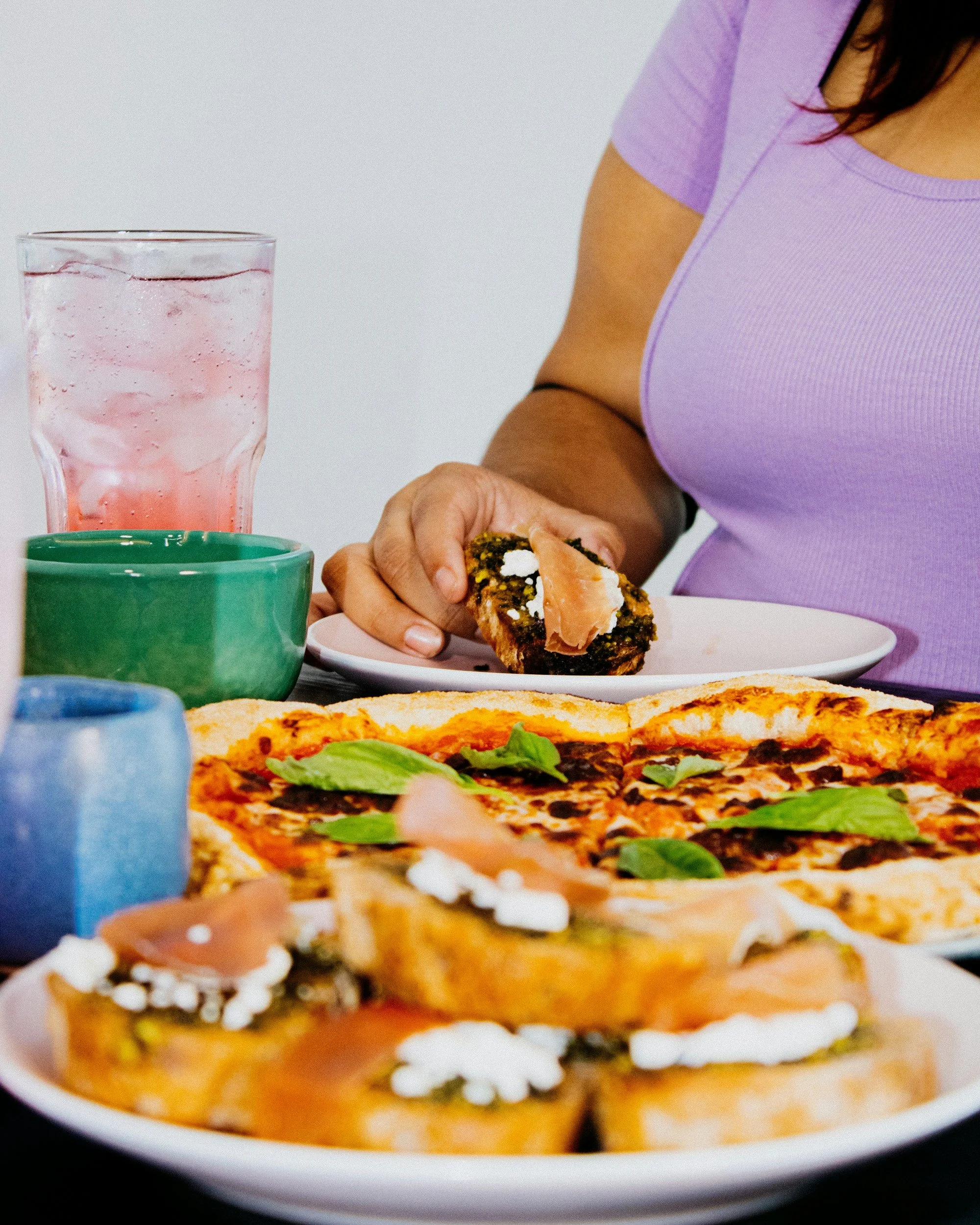 Close-up of a slice of pizza with basil leaves, cheese, and tomato sauce on a white plate. In the background, there's a glass of pink-colored drink with ice and a green cup, with a woman wearing a purple shirt holding an item in her hands.