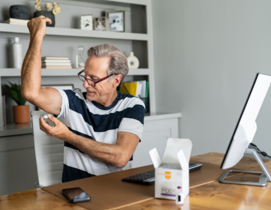 A senior man sitting at his desk flexes his bicep while preparing to attach his insulin pump, in a home office with a computer, keyboard, and shelves in the background.