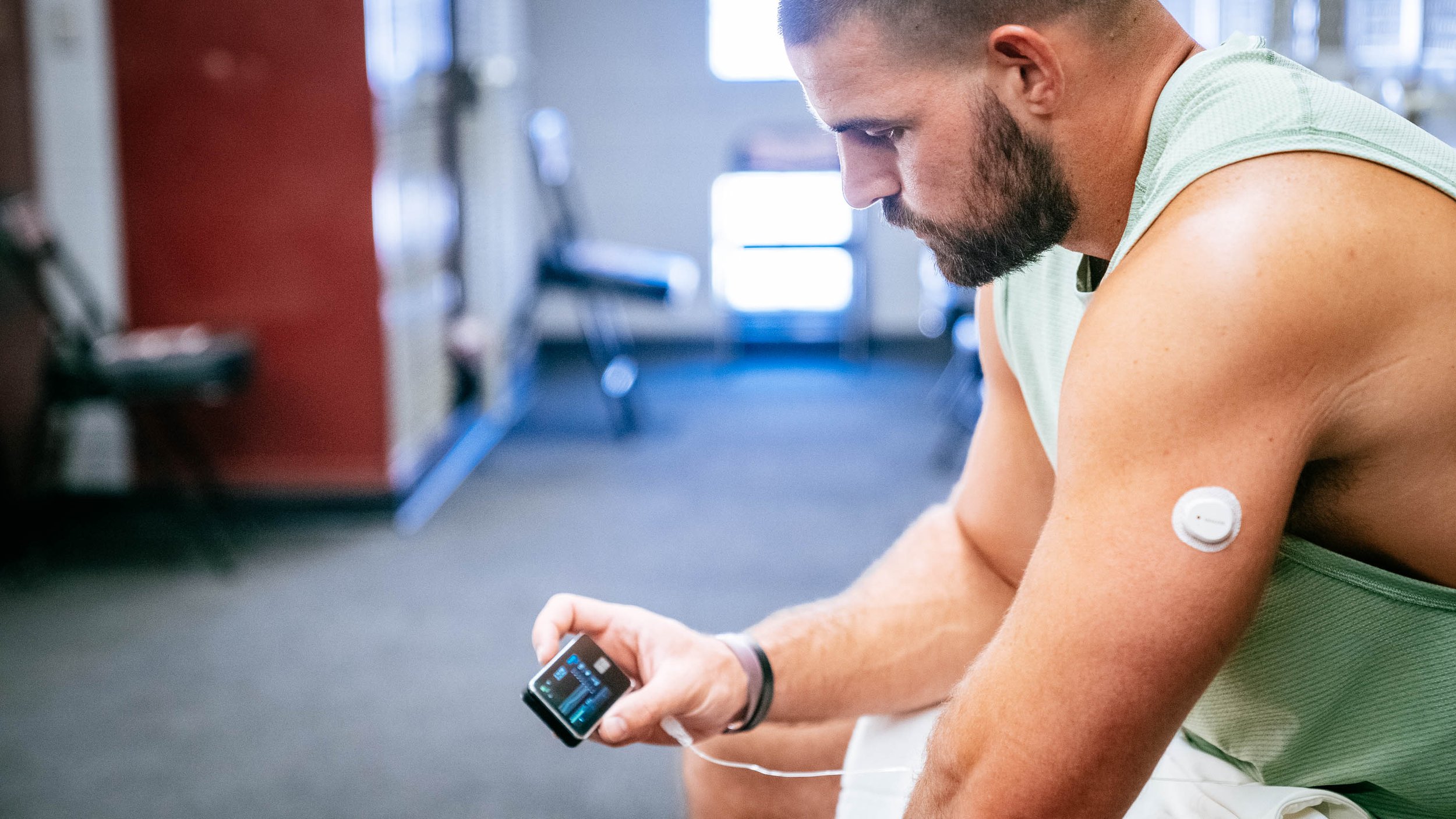 Man with a beard sitting in a gym, looking at his insulin pump with a continuous glucose monitor on his left arm.