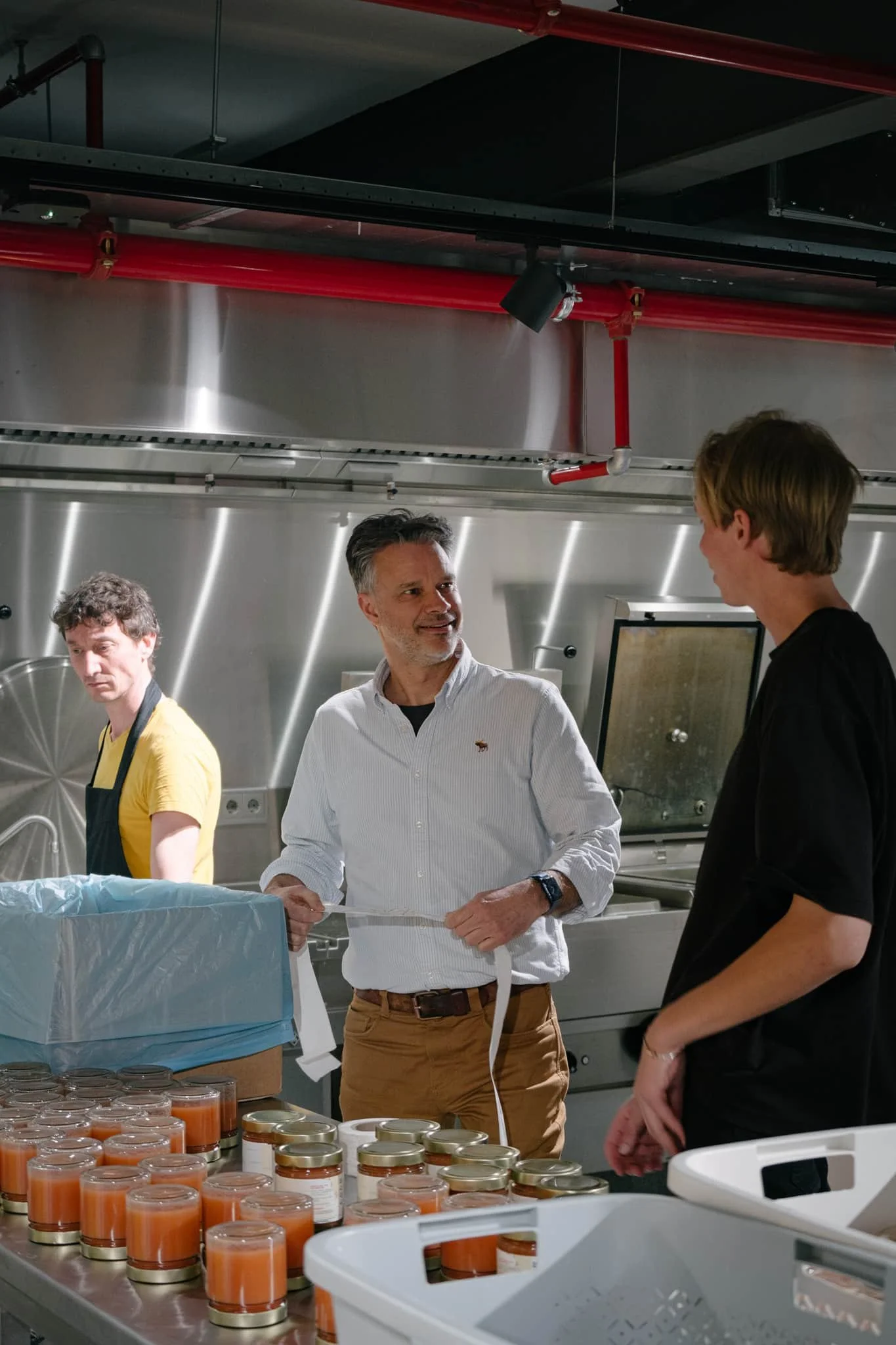 Three people are in a commercial kitchen, engaging in a conversation. One man in a white shirt and tan pants is smiling and holding a strap, speaking with a man in black. Another man in a yellow shirt and black apron is working in the background near jars of orange-colored food on the counter.