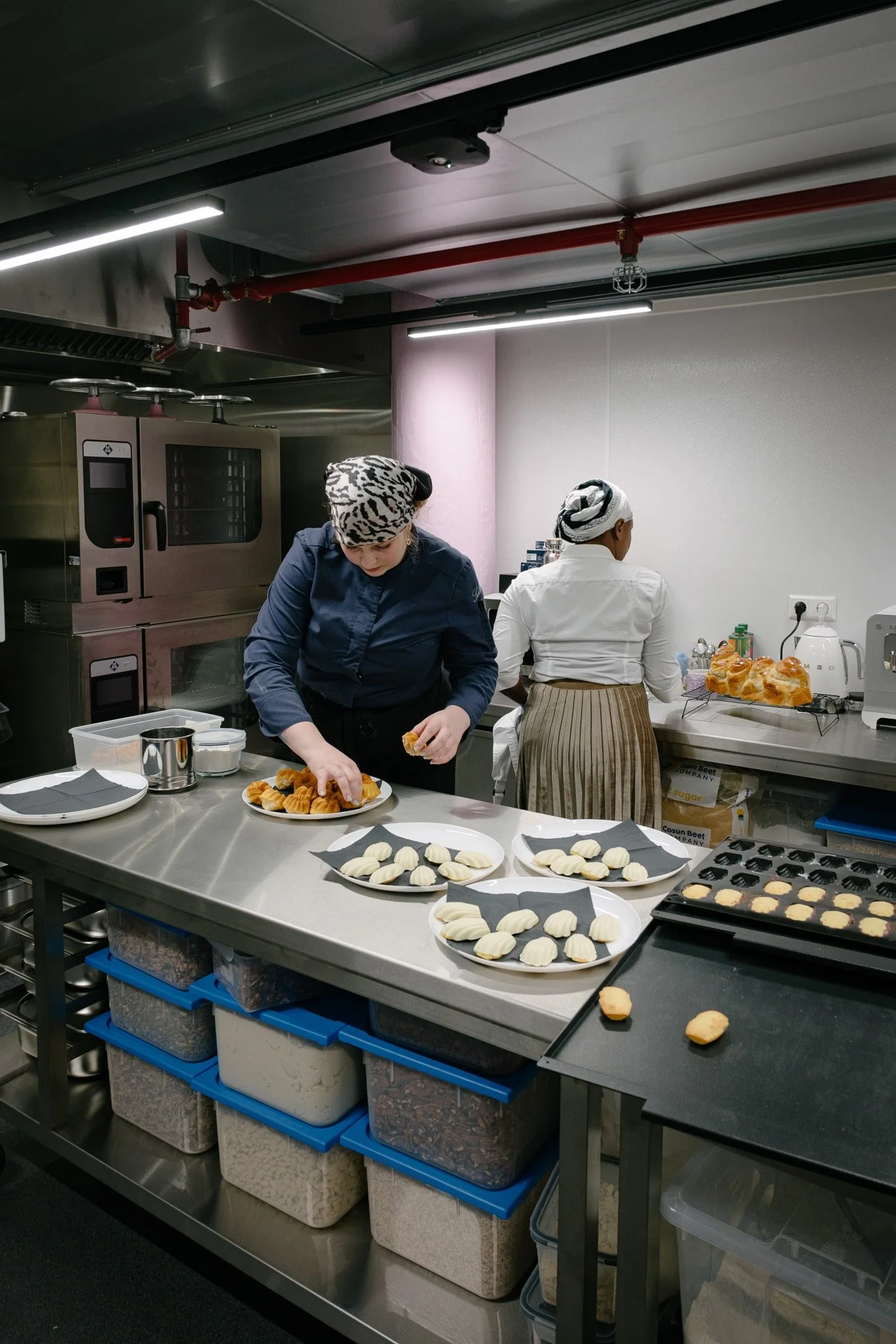 Two women working in a professional kitchen preparing pastries. One woman in navy uniform placing pastries on a white plate, while the other woman with her back facing the camera is working at the counter. The kitchen has industrial equipment and containers with ingredients underneath the counter.