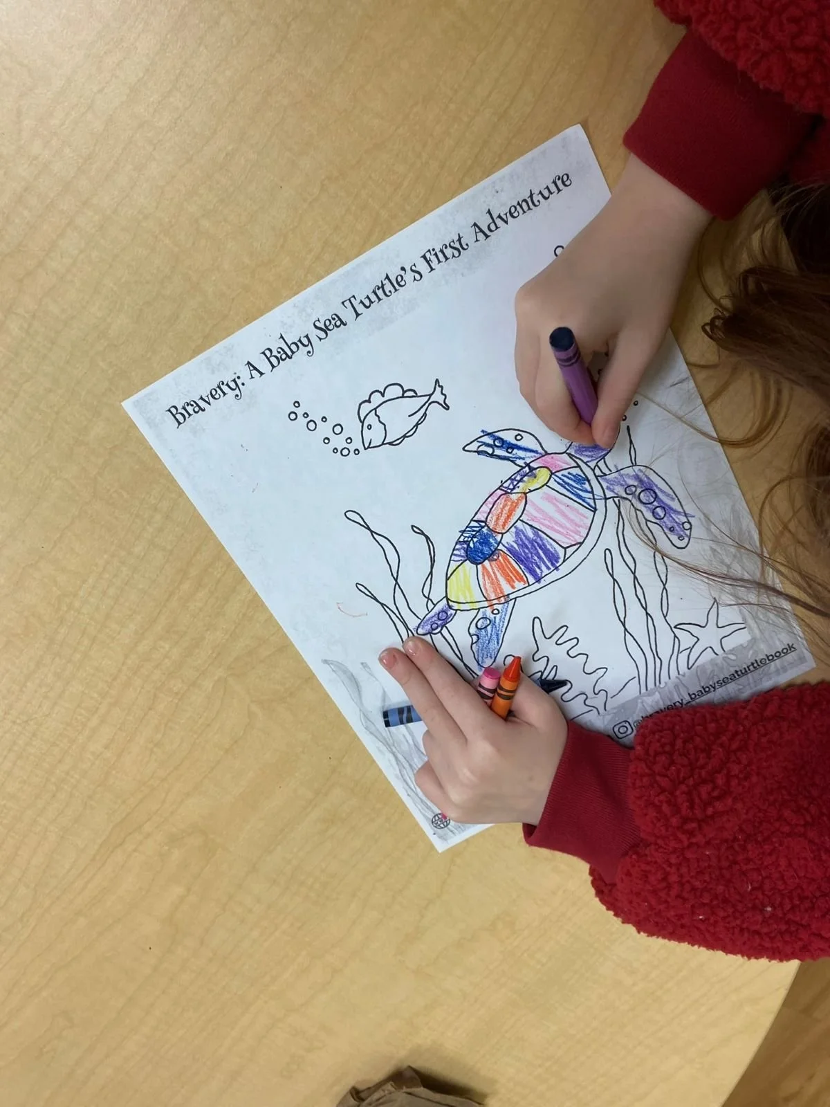 A child's hand coloring a sea turtle illustration on a worksheet titled 'Bravery: A Baby Sea Turtle's First Adventure.' The child is using crayons and coloring the turtle, which is outlined with various patterns. There is a small fish and seaweed in the illustration, and the worksheet is placed on a wooden table.