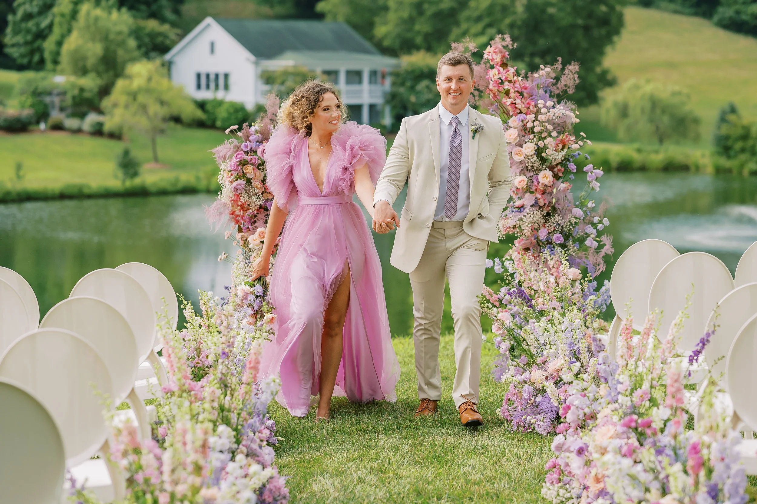 A couple walking hand in hand at an outdoor wedding ceremony by a lake. The bride has a pink wedding dress on at Mint Springs Farm. Wedding Photography by Sowing Clover Photography.