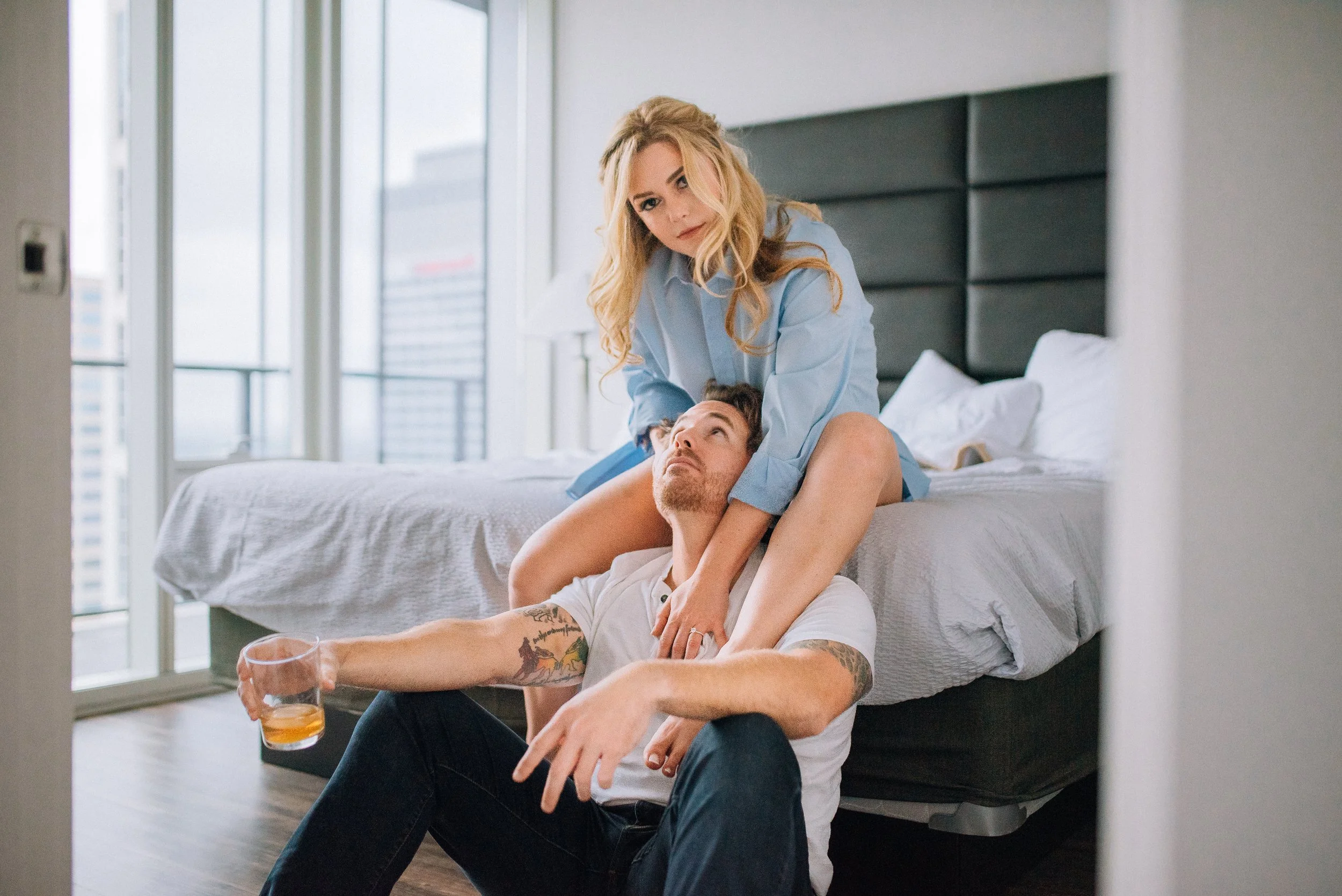 A woman sits on a bed with her legs crossed, looking at a man who is sitting on the floor leaning against the bed. The man has tattoos on his arm and is holding a glass of beer. The woman has blonde hair and is wearing a blue shirt. The room is brigh