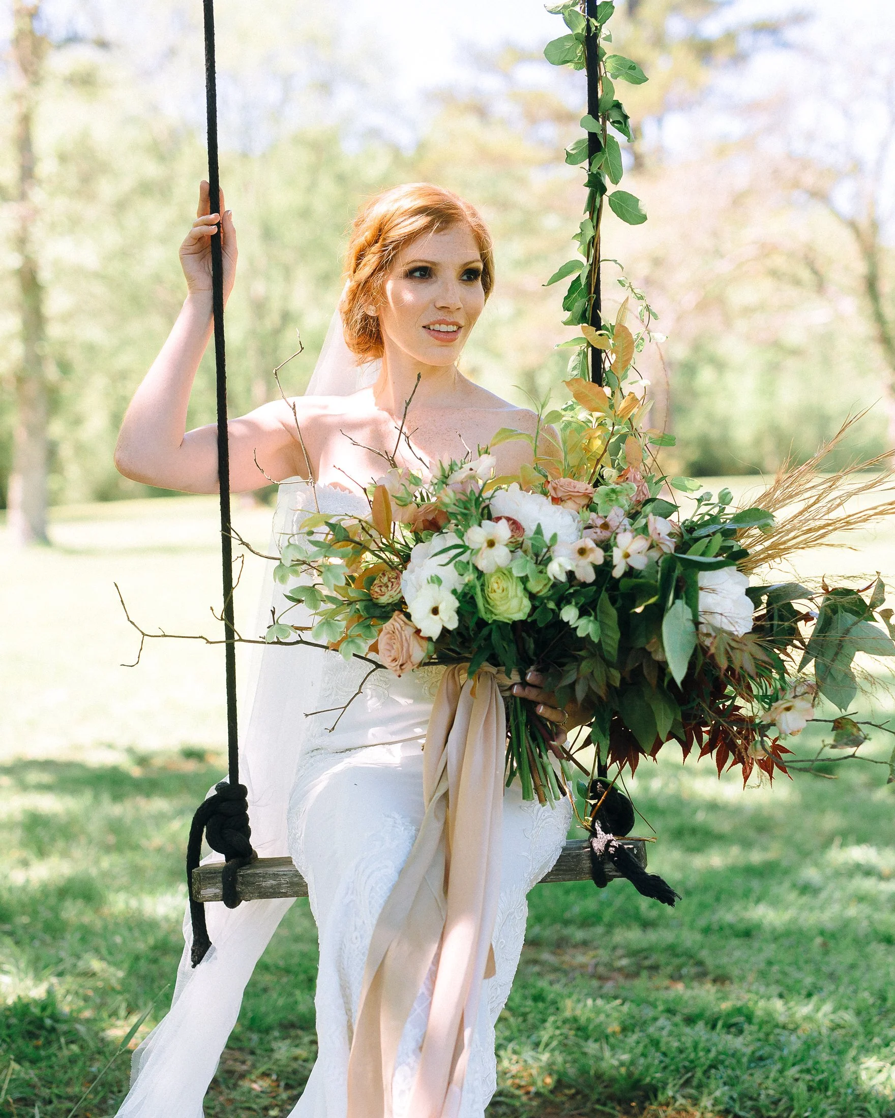 A bride with red hair in a white wedding dress sitting on a wooden swing, holding a large bouquet of flowers, outdoors at Glen Ella Springs.  Wedding Photography by Sowing Clover Photography.