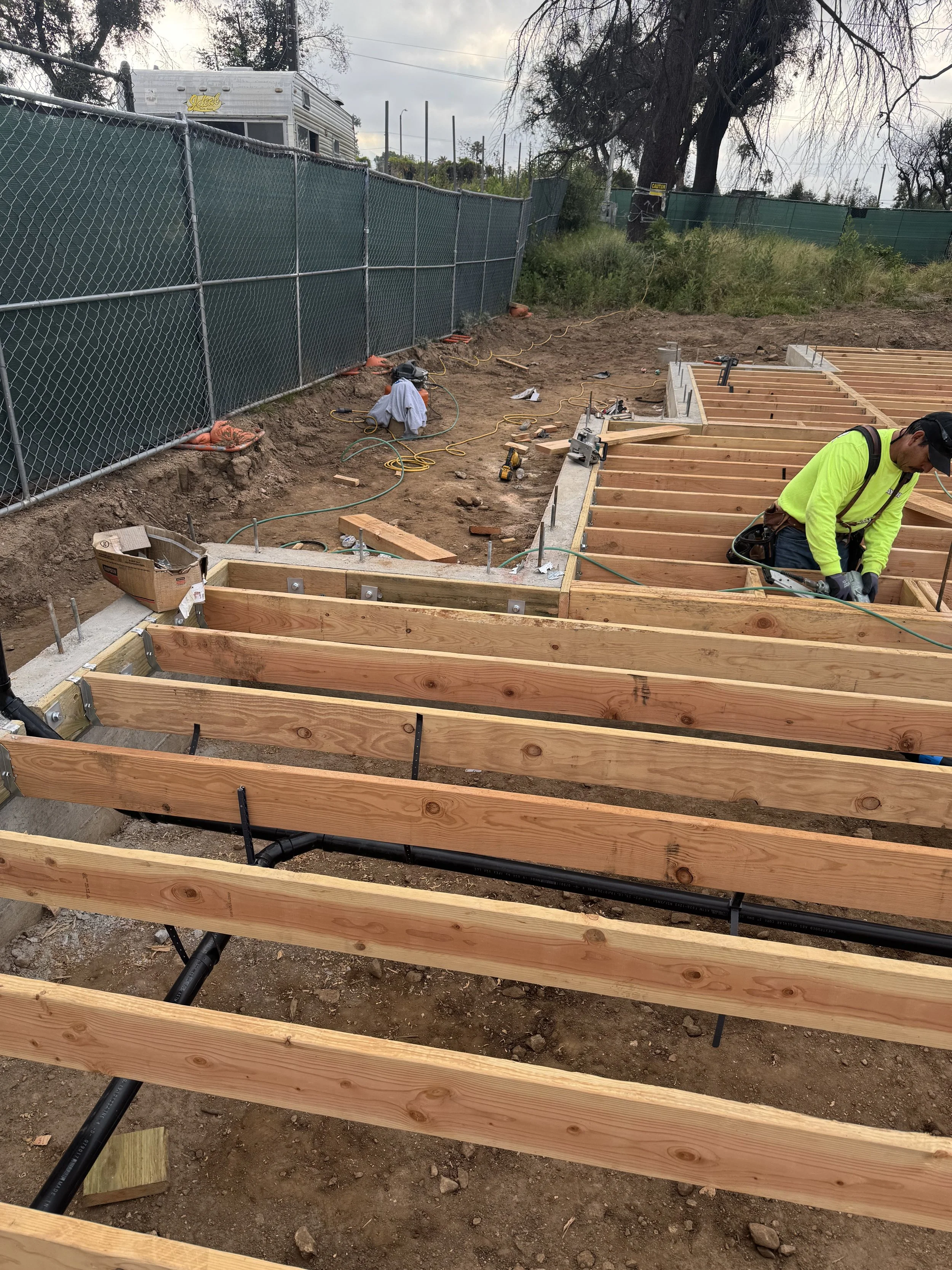 Construction workers building a wooden frame for a structure on a dirt foundation, with a chain-link fence and trees in the background.