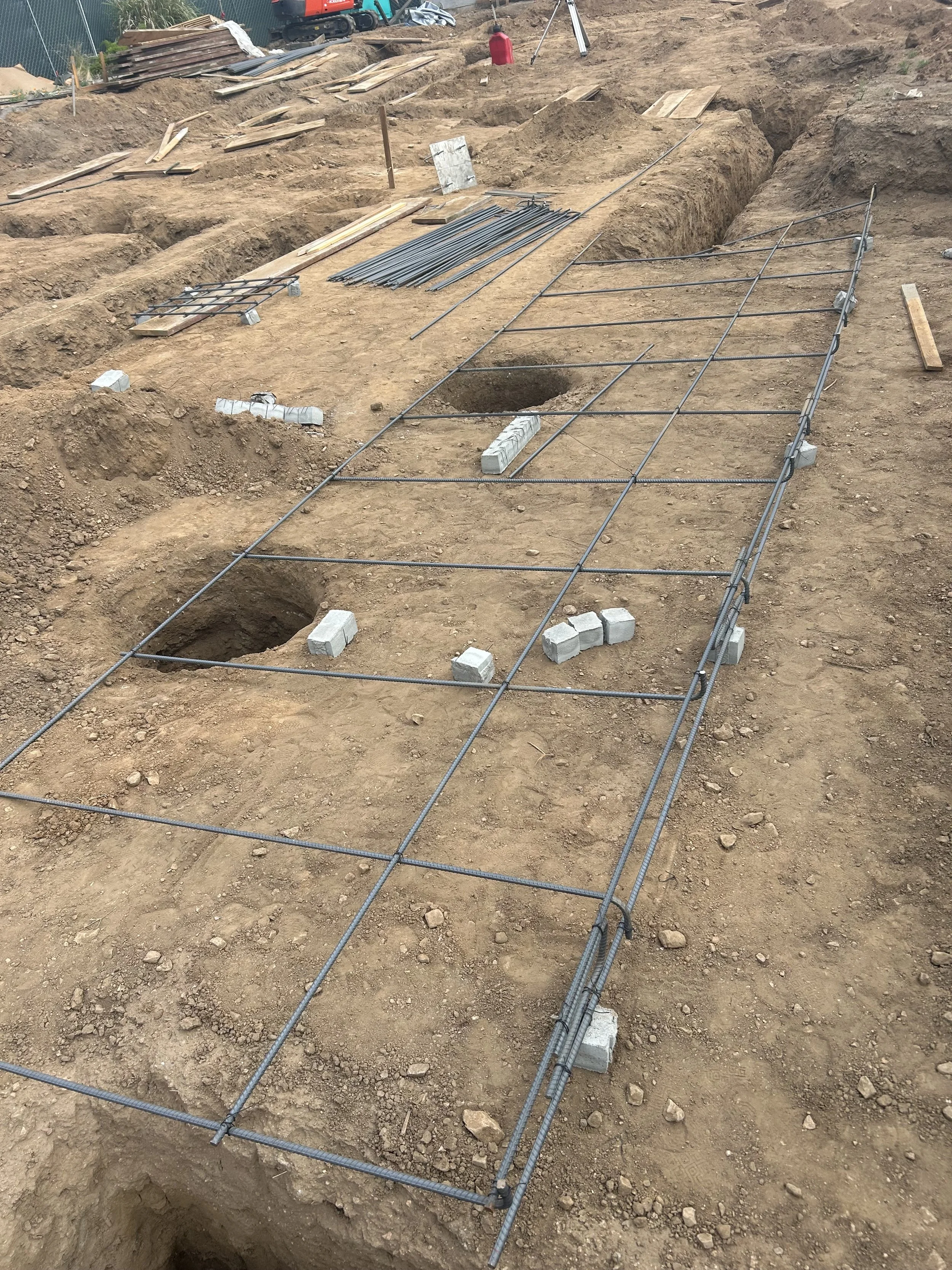 Construction site with trenches, metal rebar grid, concrete blocks, and dirt, indicating foundation work in progress.