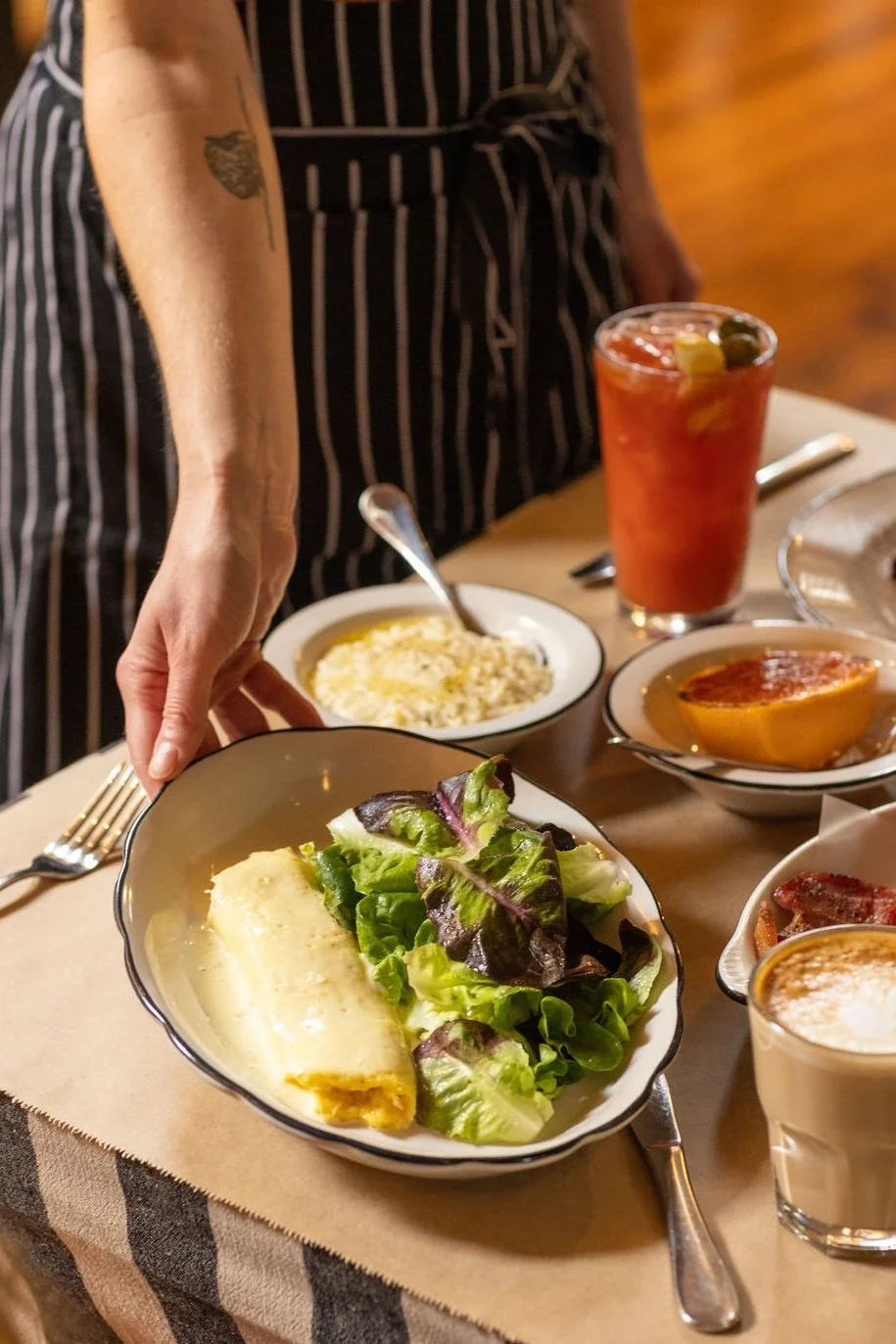 A person in a black and white striped apron setting a table with dishes including a salad with lettuce and a rolled egg dish, a bowl of rice, a bowl with a half grapefruit, and a glass of Bloody Mary with olives and lemon slices.