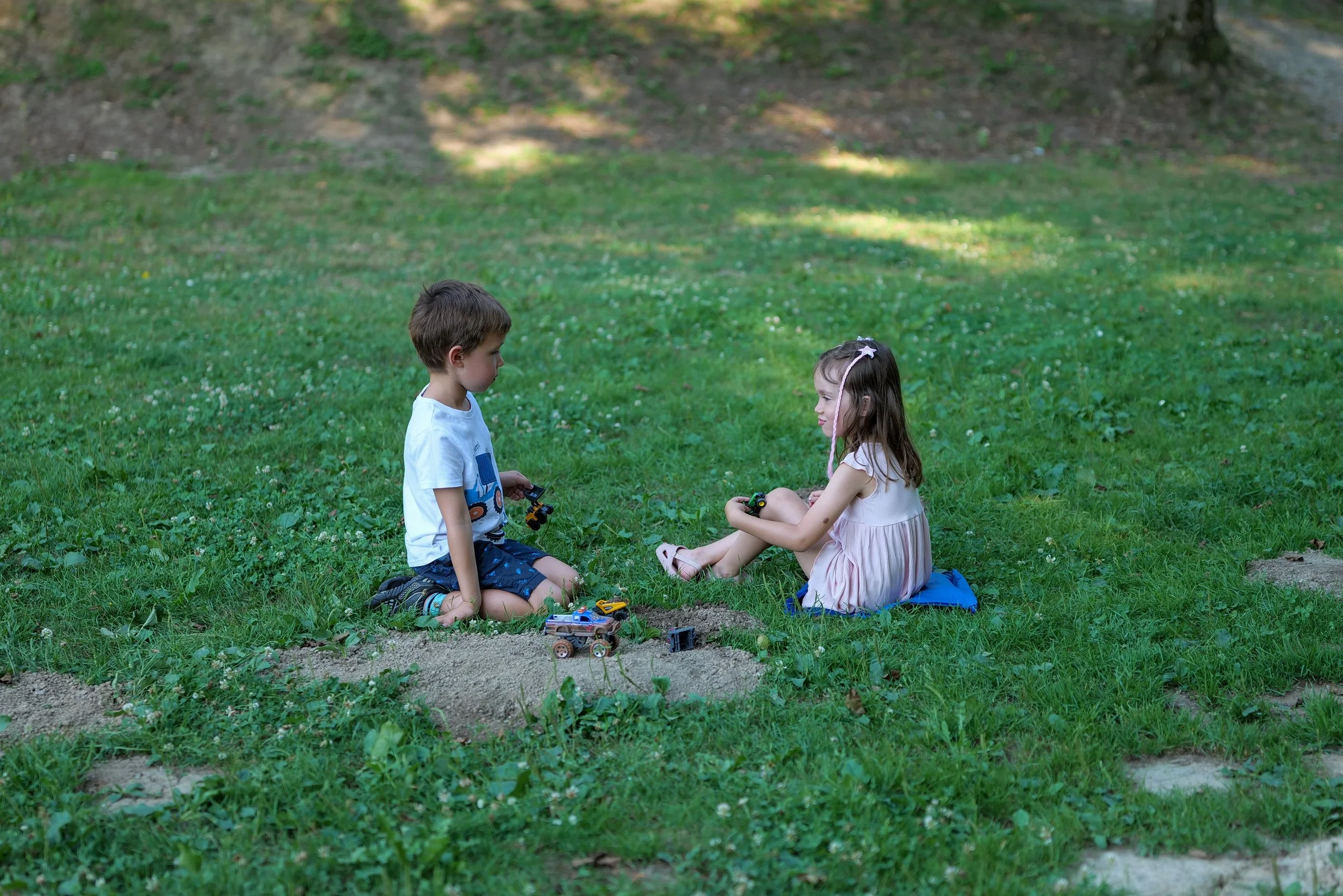 A young boy and girl sitting on the grass in a park, playing together with toy trucks and other small toys.
