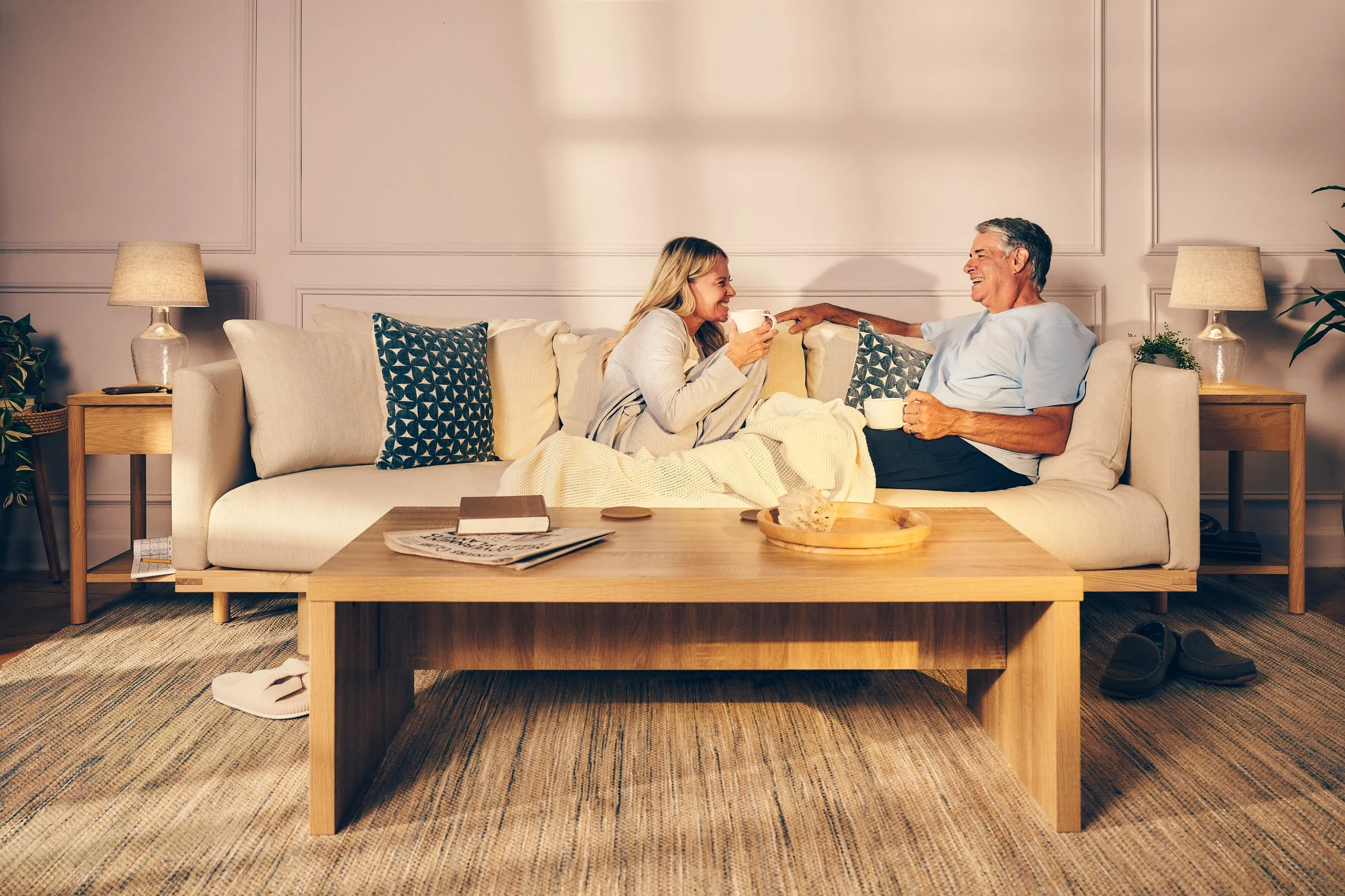 A middle-aged man and a young woman sitting on a cream-colored sofa, smiling and enjoying a conversation while holding coffee mugs. The man is wearing a light blue shirt, and the woman is in a light-colored sweater. The living room is warmly lit with two side tables and lamps, patterned pillows, a wooden coffee table, a framed wall, and a cozy atmosphere.