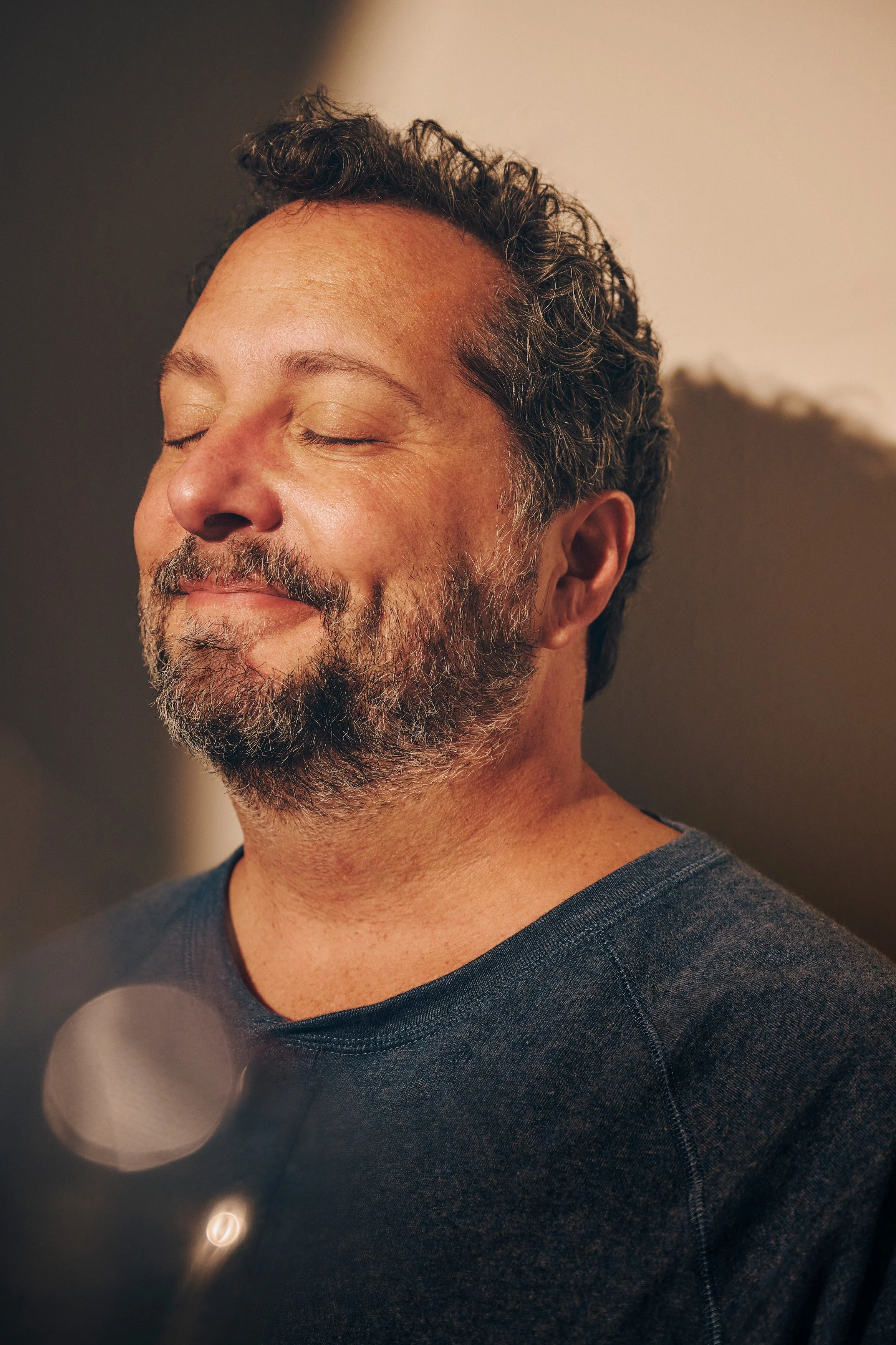 A middle-aged man with a beard and short curly hair, smiling with his eyes closed, standing against a neutral background.