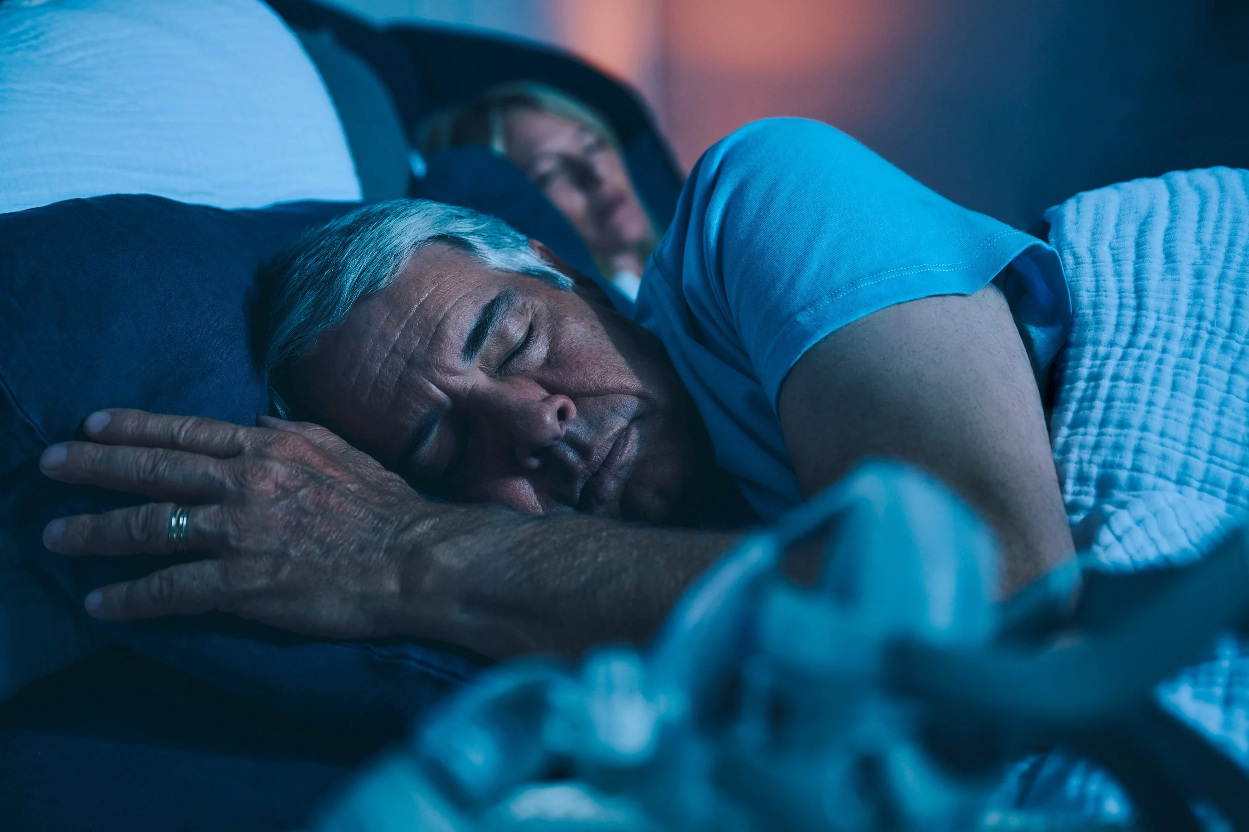 An elderly man sleeping peacefully on a bed, with another person blurred in the background.