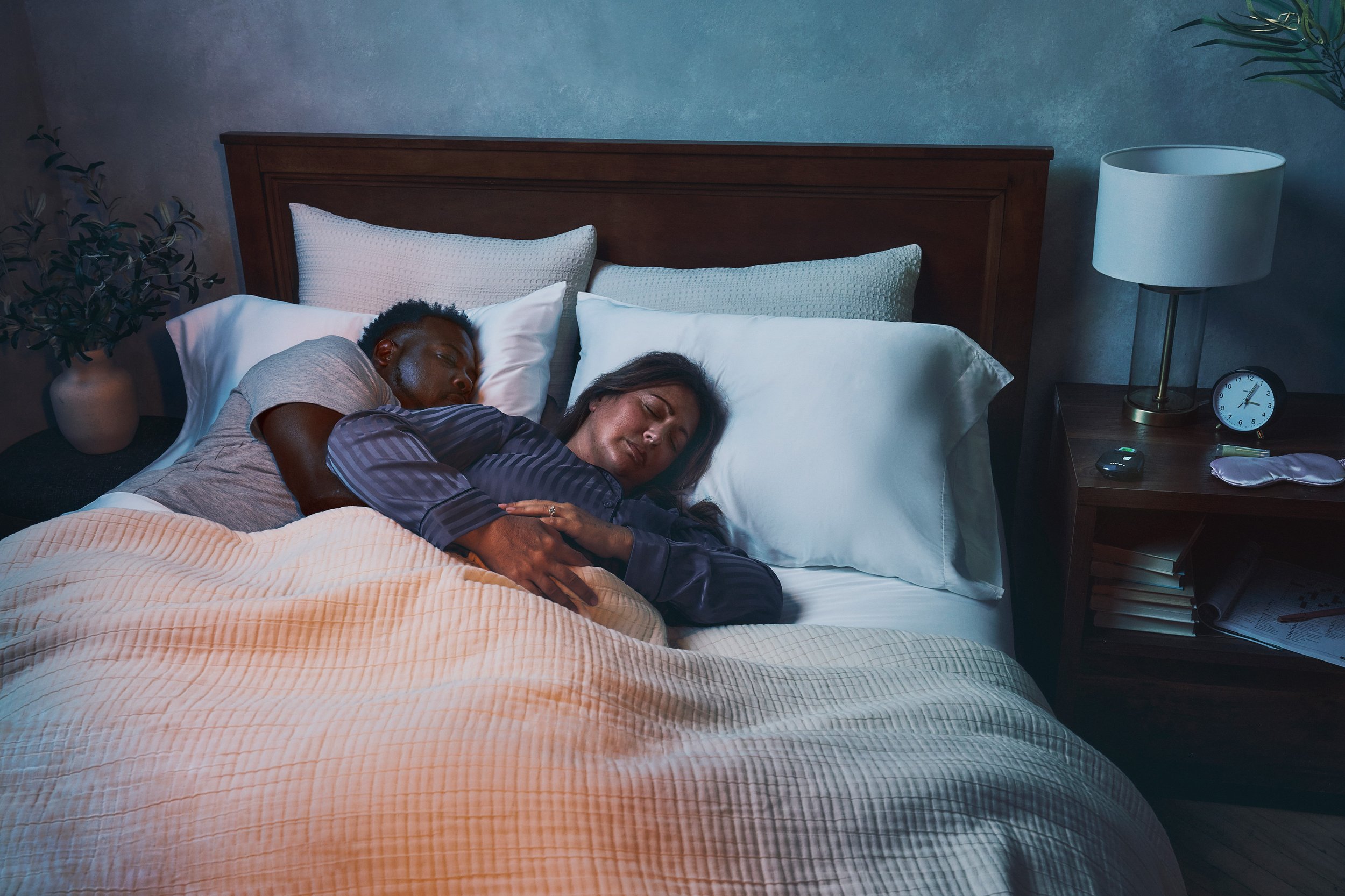 A couple sleeping together in bed in a bedroom at night, with a nightstand, a clock, a lamp, and plants nearby.
