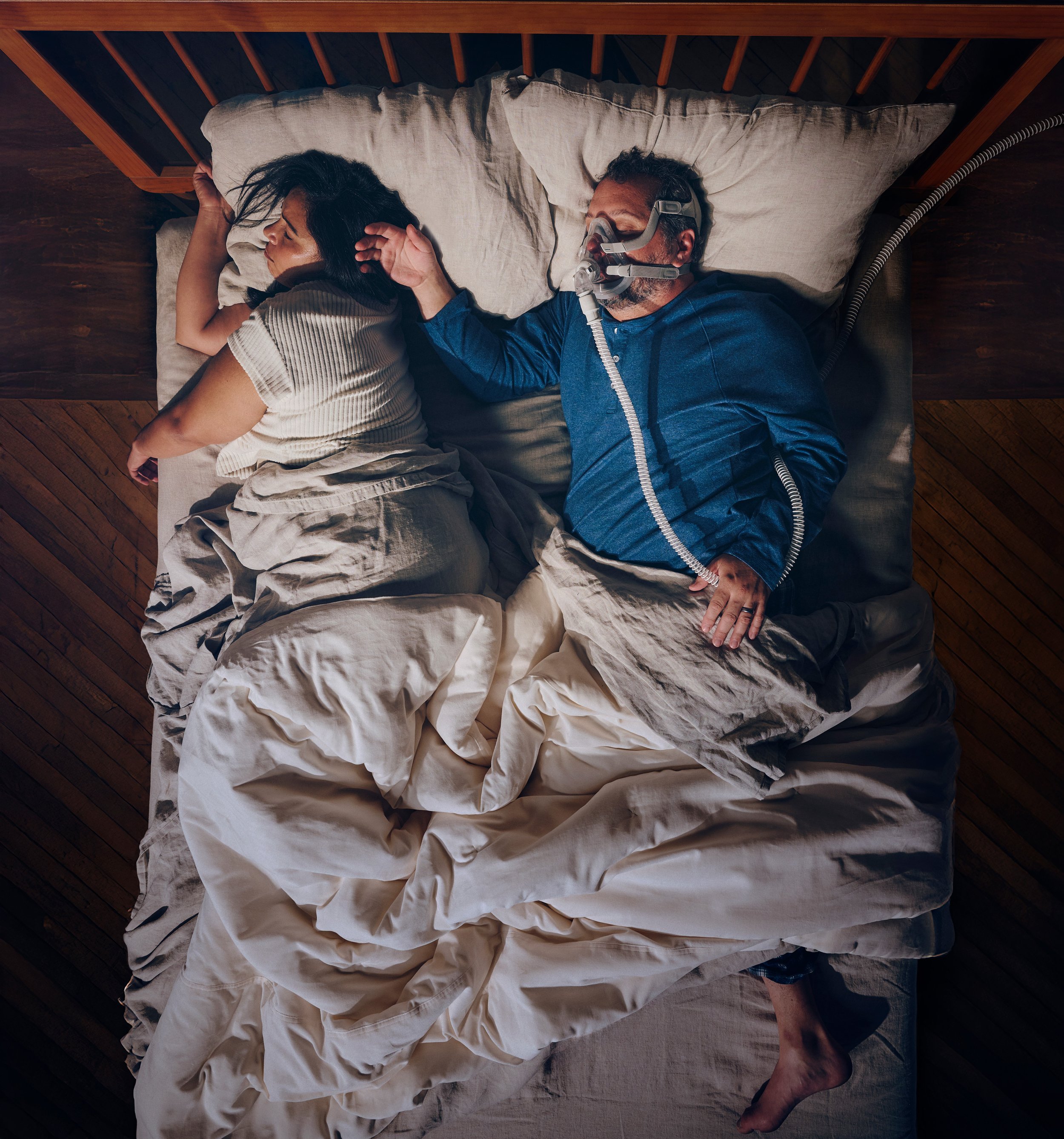 An overhead view of a couple sleeping in bed. The woman is on her side, with dark hair, wearing a beige top. The man is lying on his back, wearing a blue shirt, and using a CPAP mask.