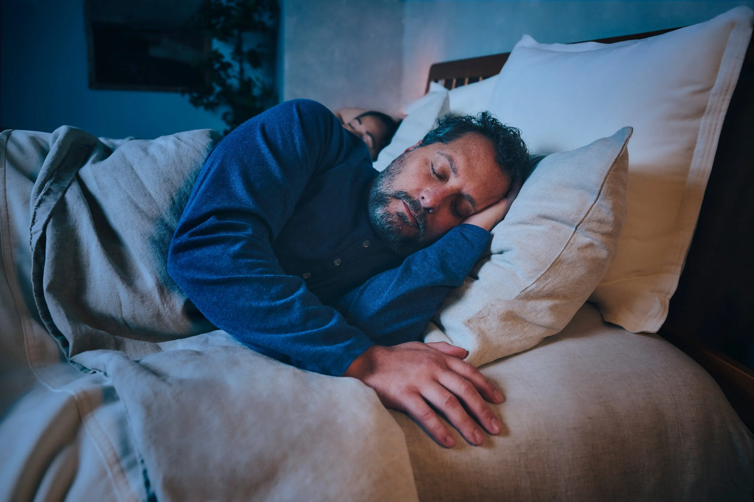 A man with curly hair and a beard sleeping on his side in bed, resting his head on a pillow. Another person is slightly visible in the background, also sleeping.