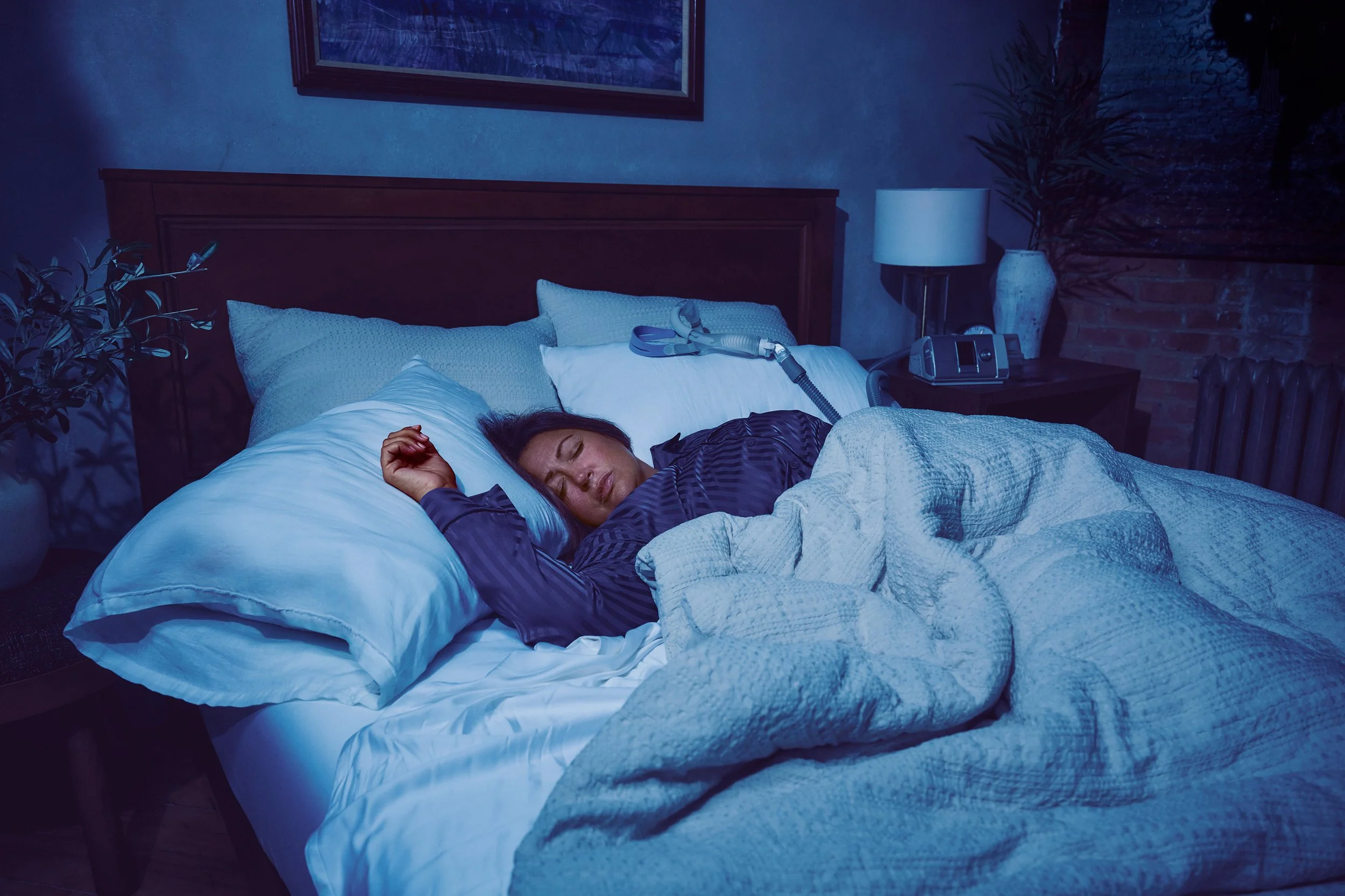 Woman sleeping in bed with a CPAP machine beside her in a dimly lit bedroom.