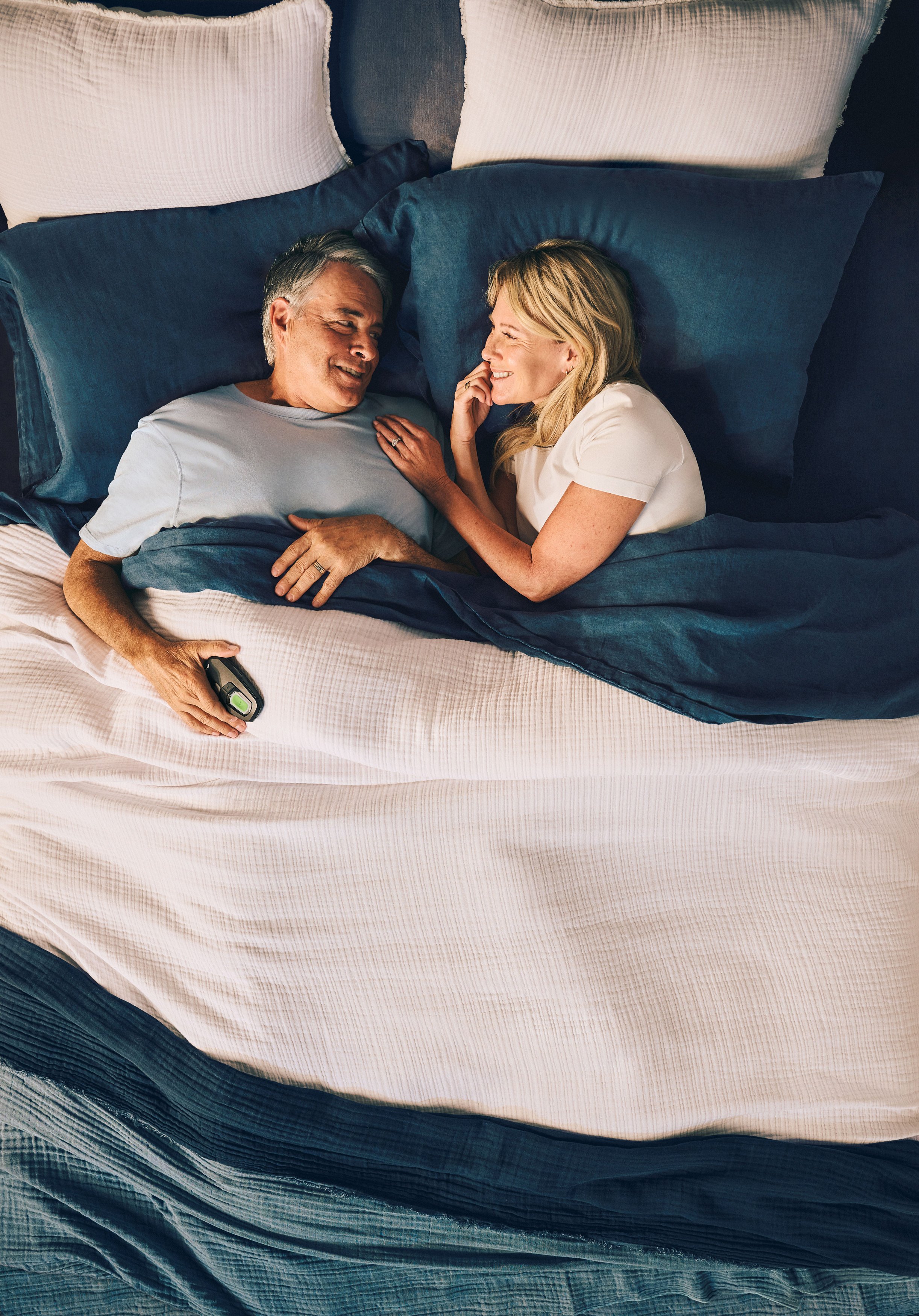 A man and woman smiling and talking in bed with blue and white bedding and pillows.