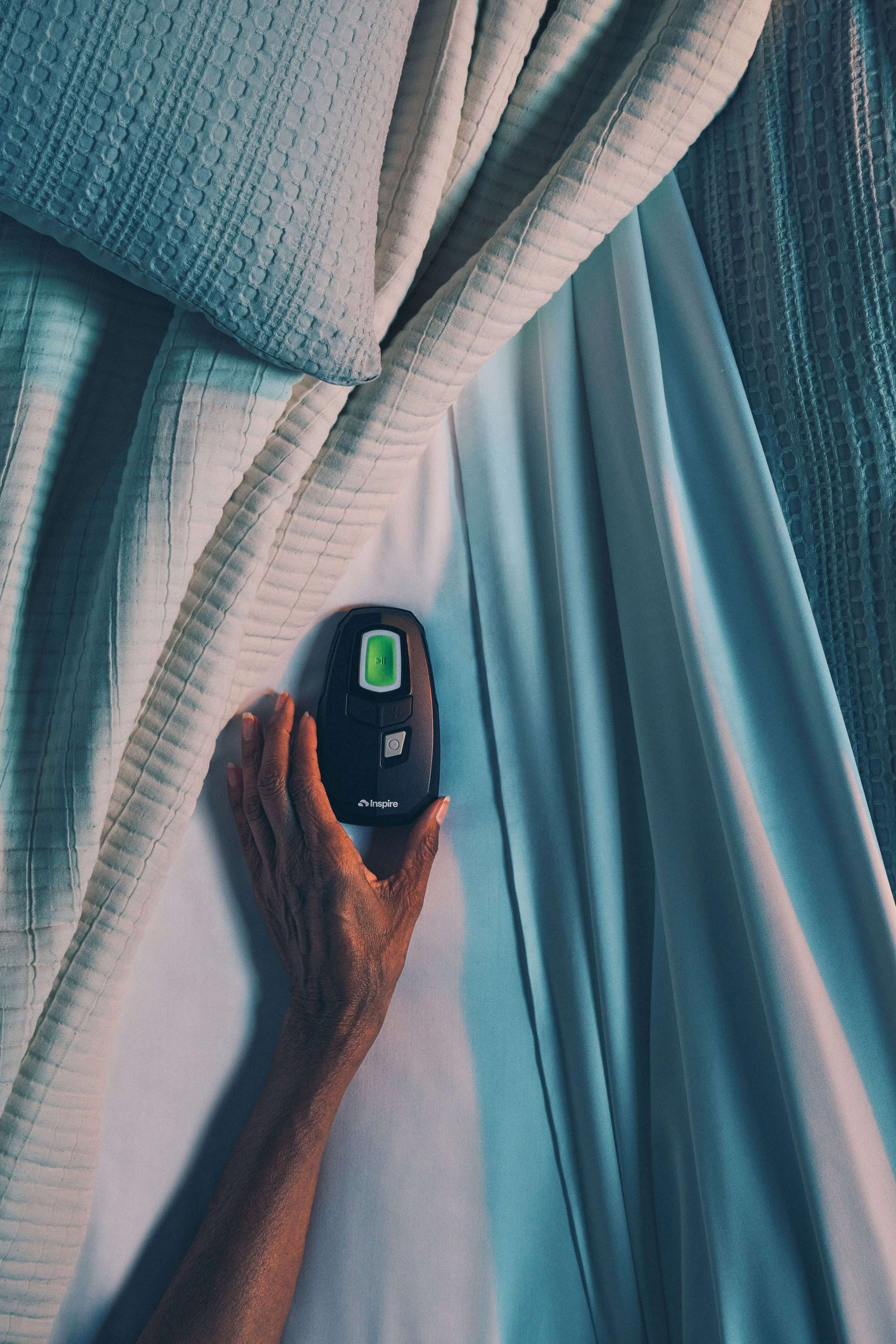 A hand adjusts a bed remote control on a bed with white sheets and pillows.