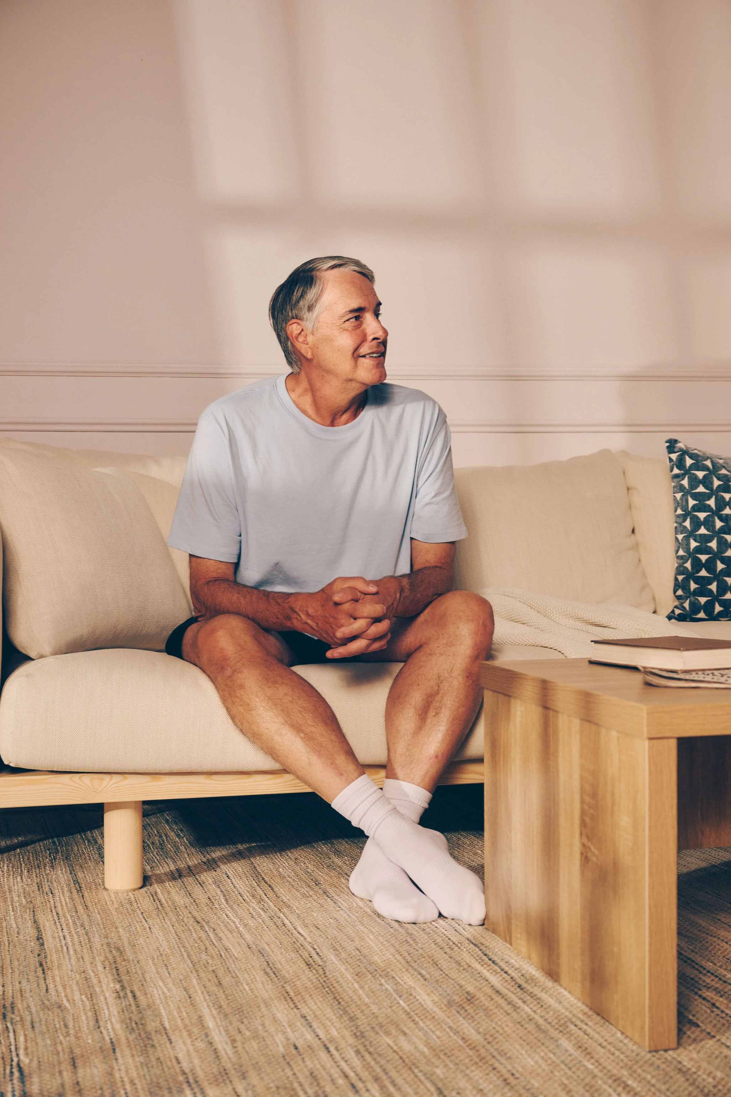Older man sitting on a beige sofa, wearing a light blue t-shirt, black shorts, and white socks, with sunlight casting shadows on the wall behind.