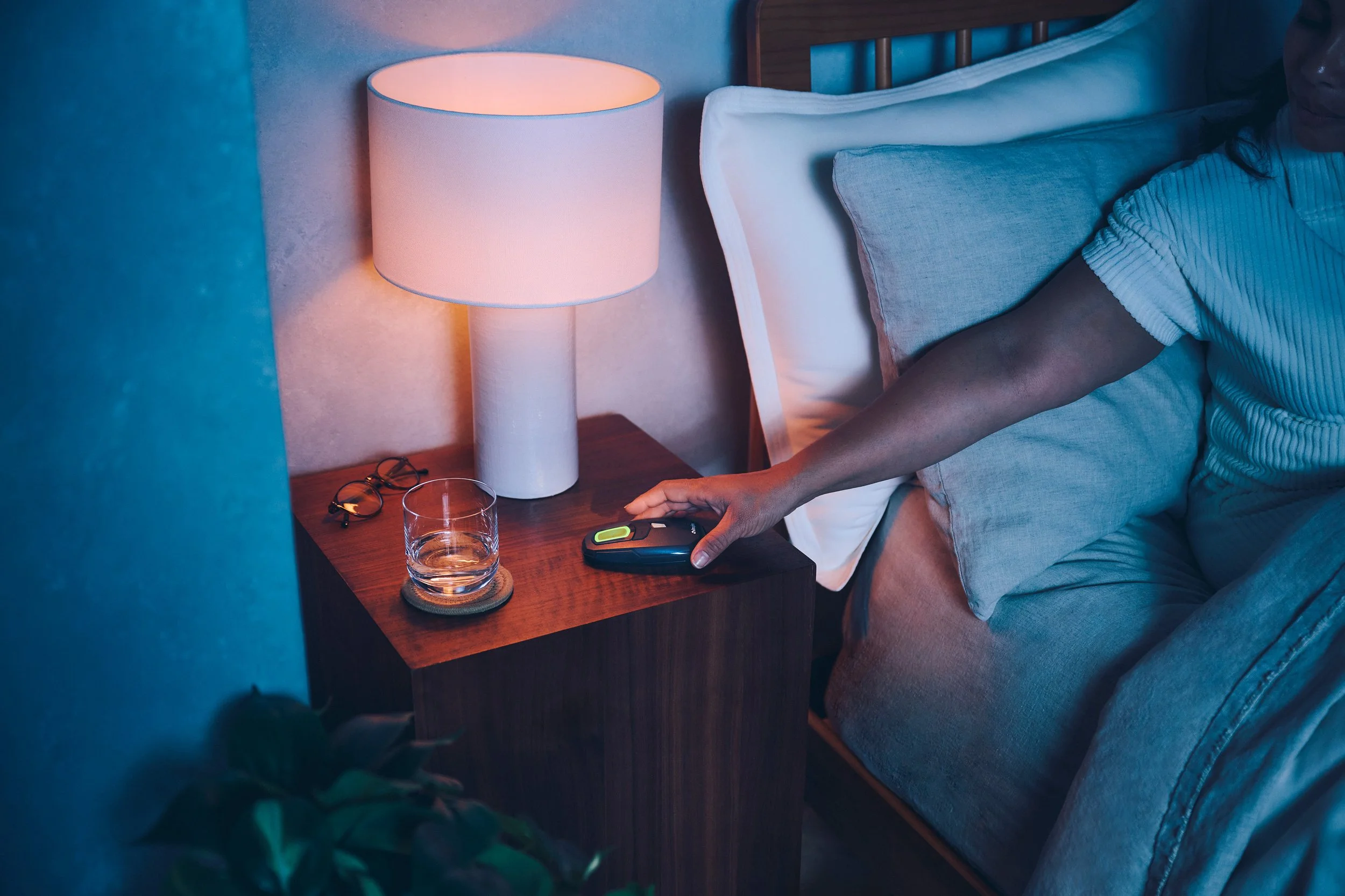 A person reaches for a TV remote on a wooden nightstand, next to a glass of water and a pair of glasses, in a cozy bedroom with a bedside lamp.