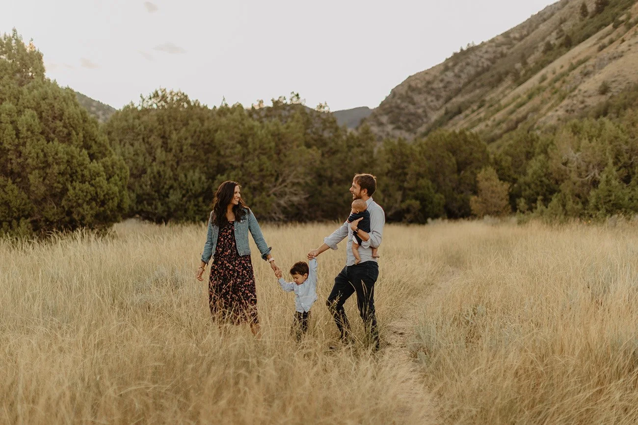 Family of four walking hand-in-hand through a grassy field with mountains and trees in the background.