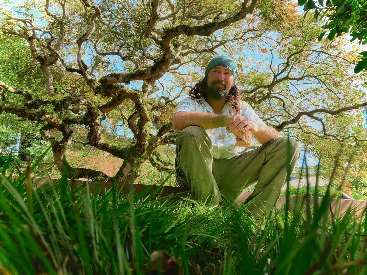 A man with long hair and a beard wearing a blue bandana, sitting cross-legged on the grass under a large, twisted tree with green foliage. He is holding hands together and smiling at the camera. The photo is taken from a low angle, showing the vibran