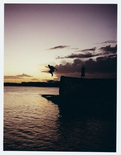 Silhouette of a person jumping off a pier into the water with another person standing on the pier at sunset, with clouds and a calm body of water in the background.