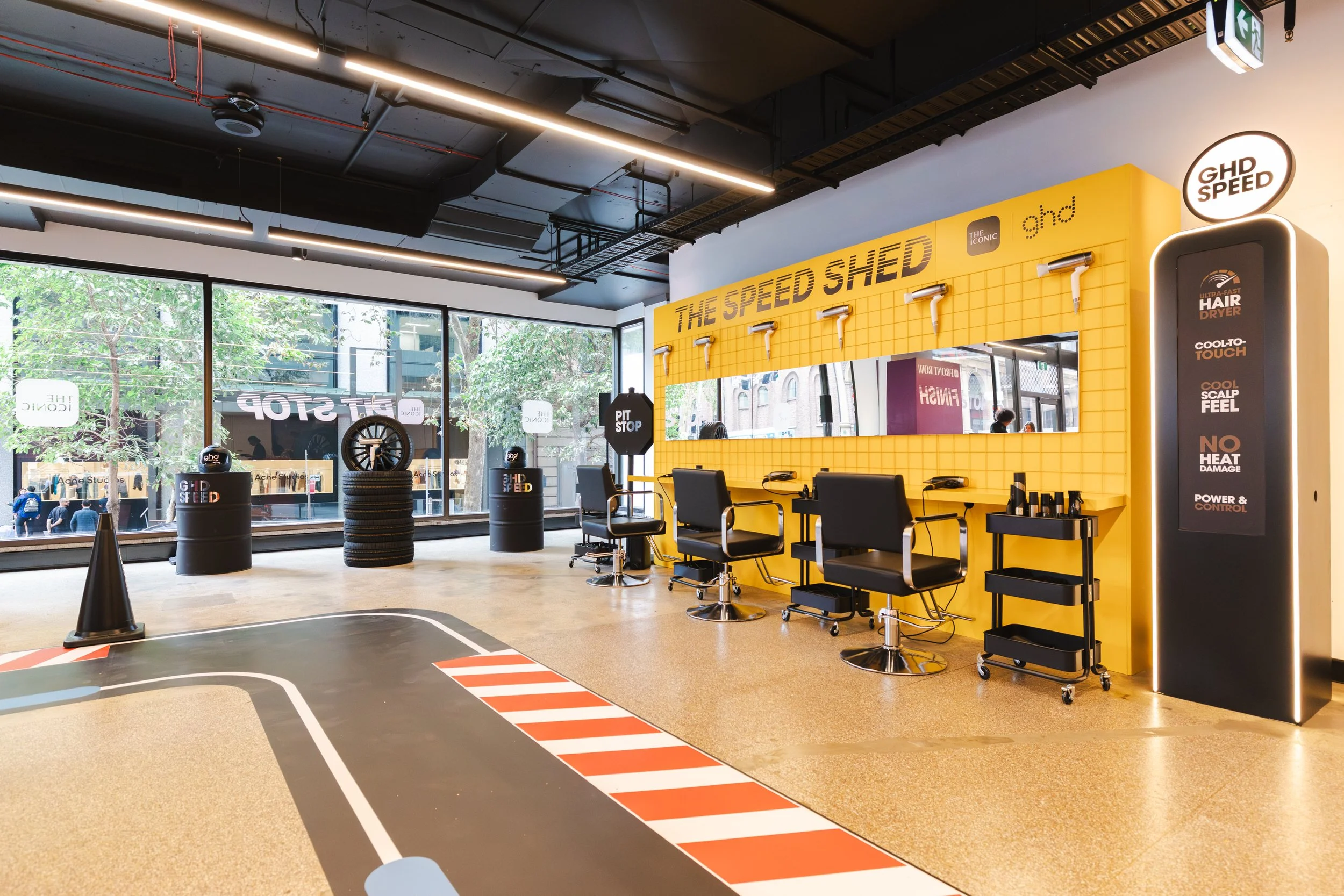 A bright, modern interior of a hair salon with a yellow wall, several styling chairs, hair dryers, and a large window showing a city street outside.