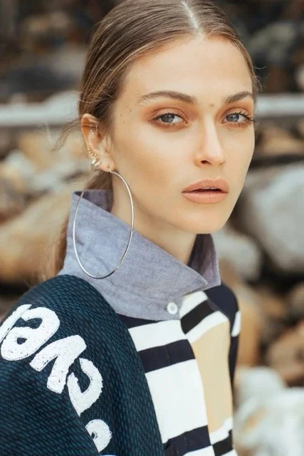 A young woman with light hair tied back, wearing a large hoop earring, a blue and white striped shirt under a gray collared shirt, and holding a navy blue bag, standing outdoors with logs in the background.