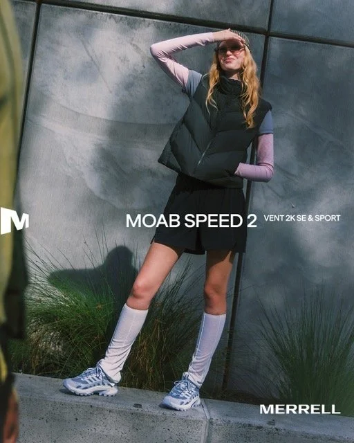 A woman with long blonde hair in sunglasses, wearing a black puffer vest, purple long sleeve shirt, black shorts, white knee-high socks, and running shoes, standing outdoors against a concrete wall, striking a pose with one hand on her forehead and o